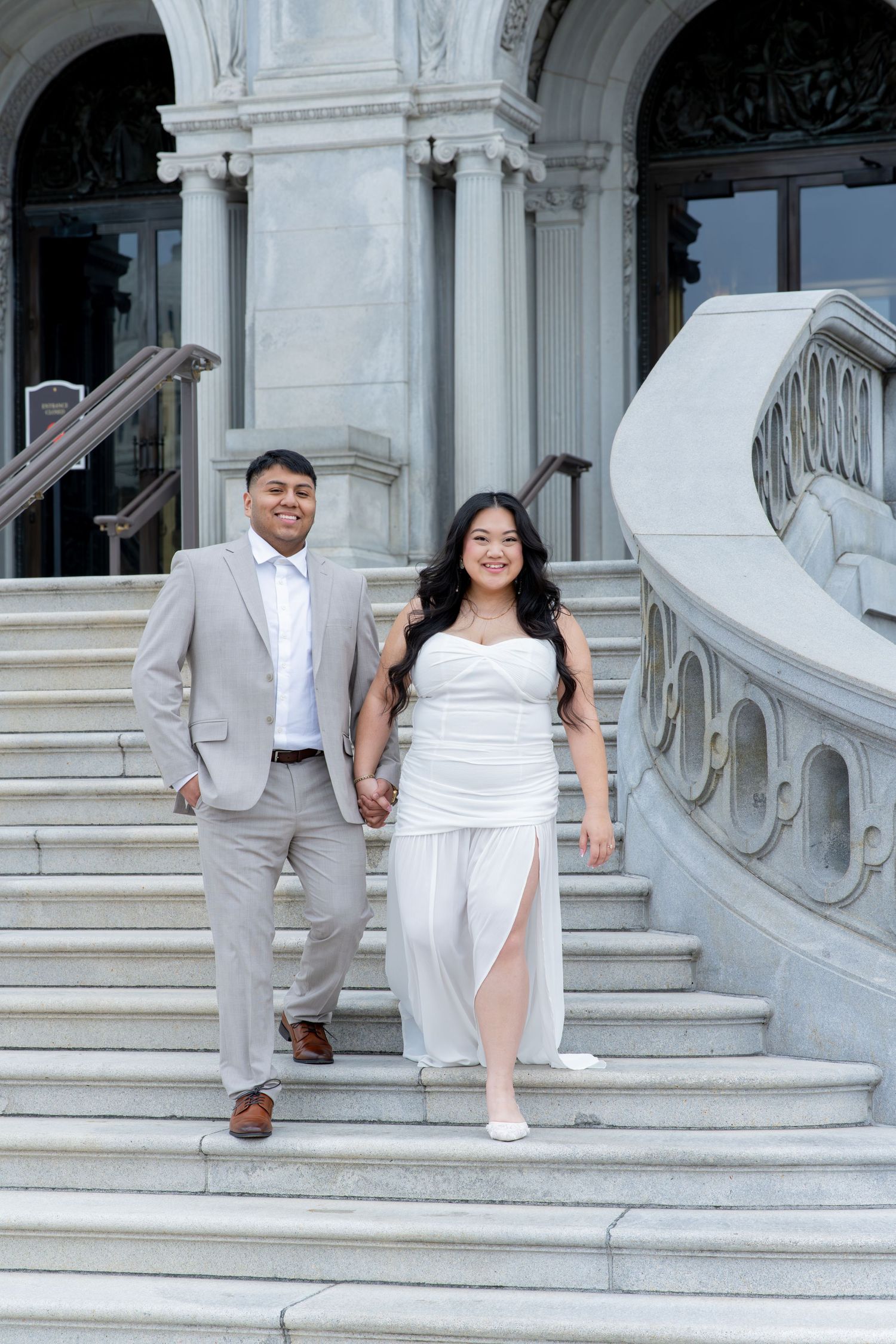 Elegant couple in wedding attire walking down marble steps of a grand neoclassical building.