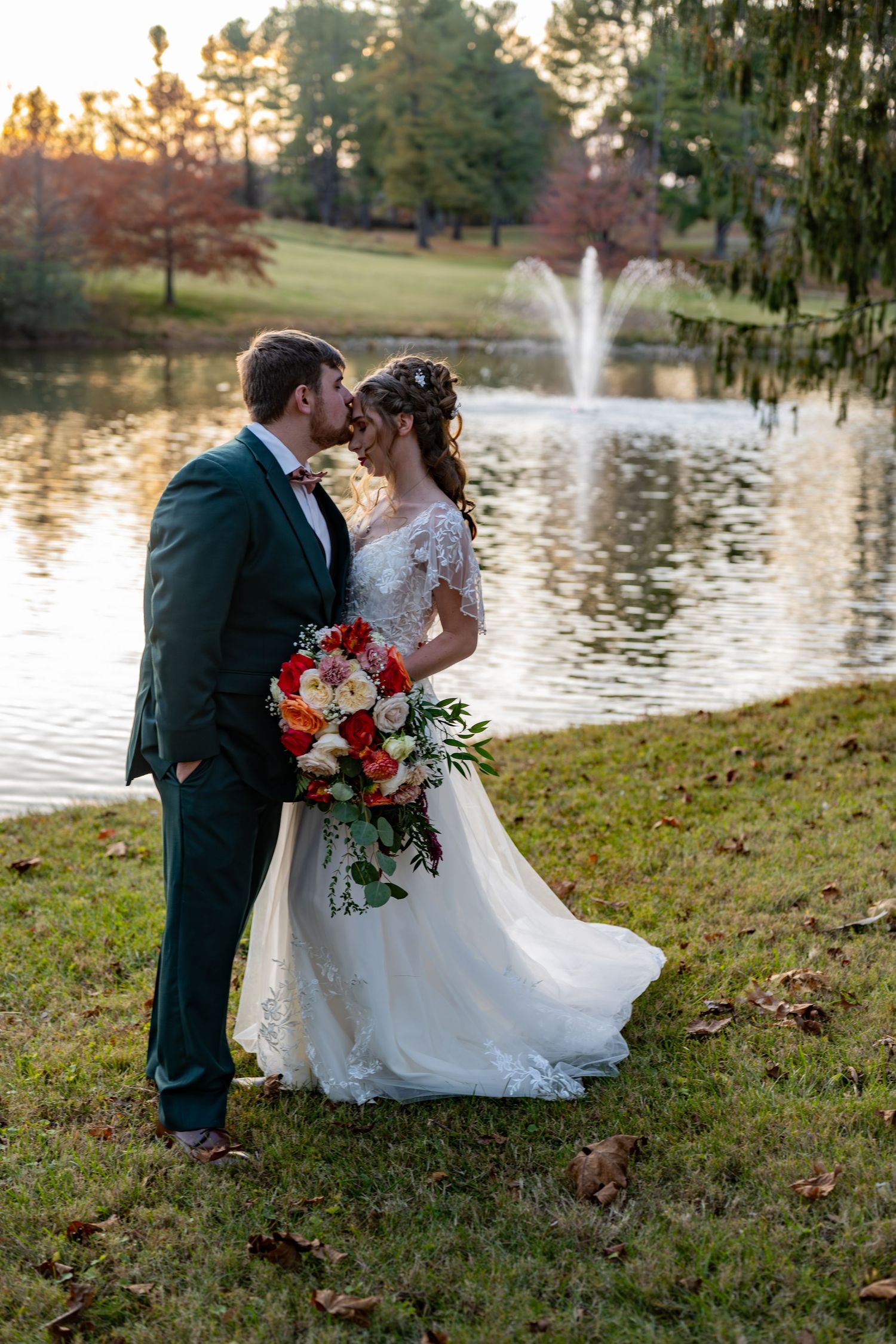 A romantic wedding couple sits on grassy hill by a lake with fountain at sunset in autumn.