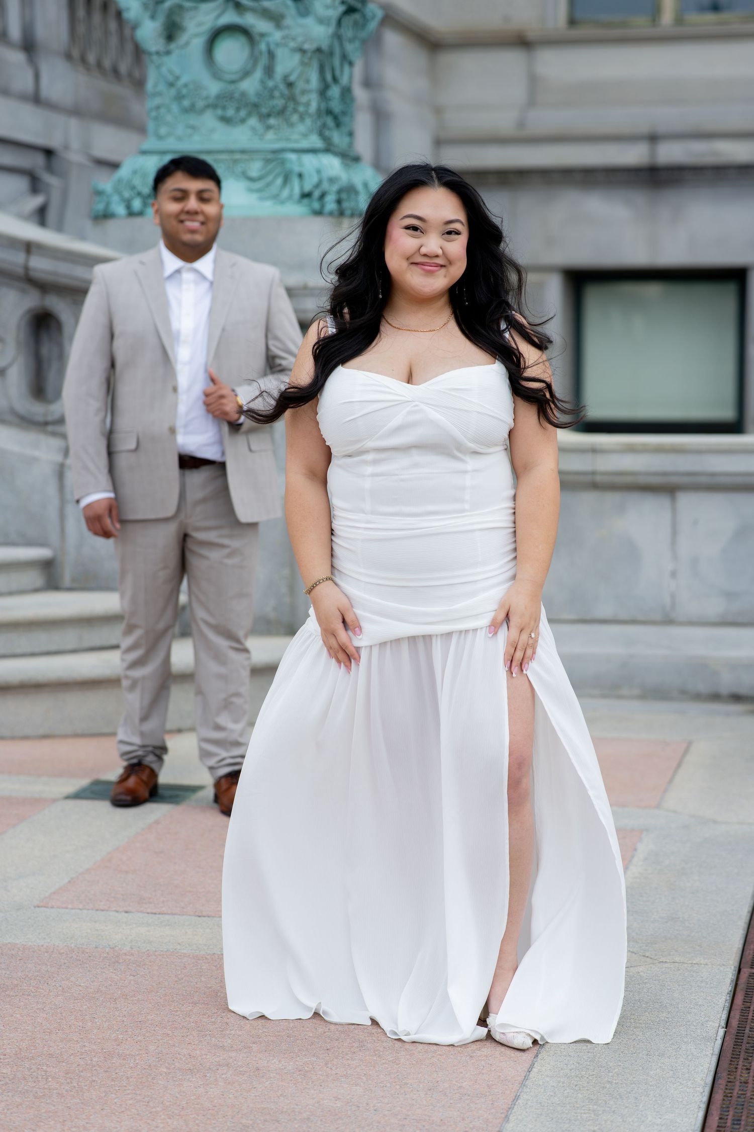 Bride in white strapless gown smiles as groom in tan suit poses behind her on stone steps.