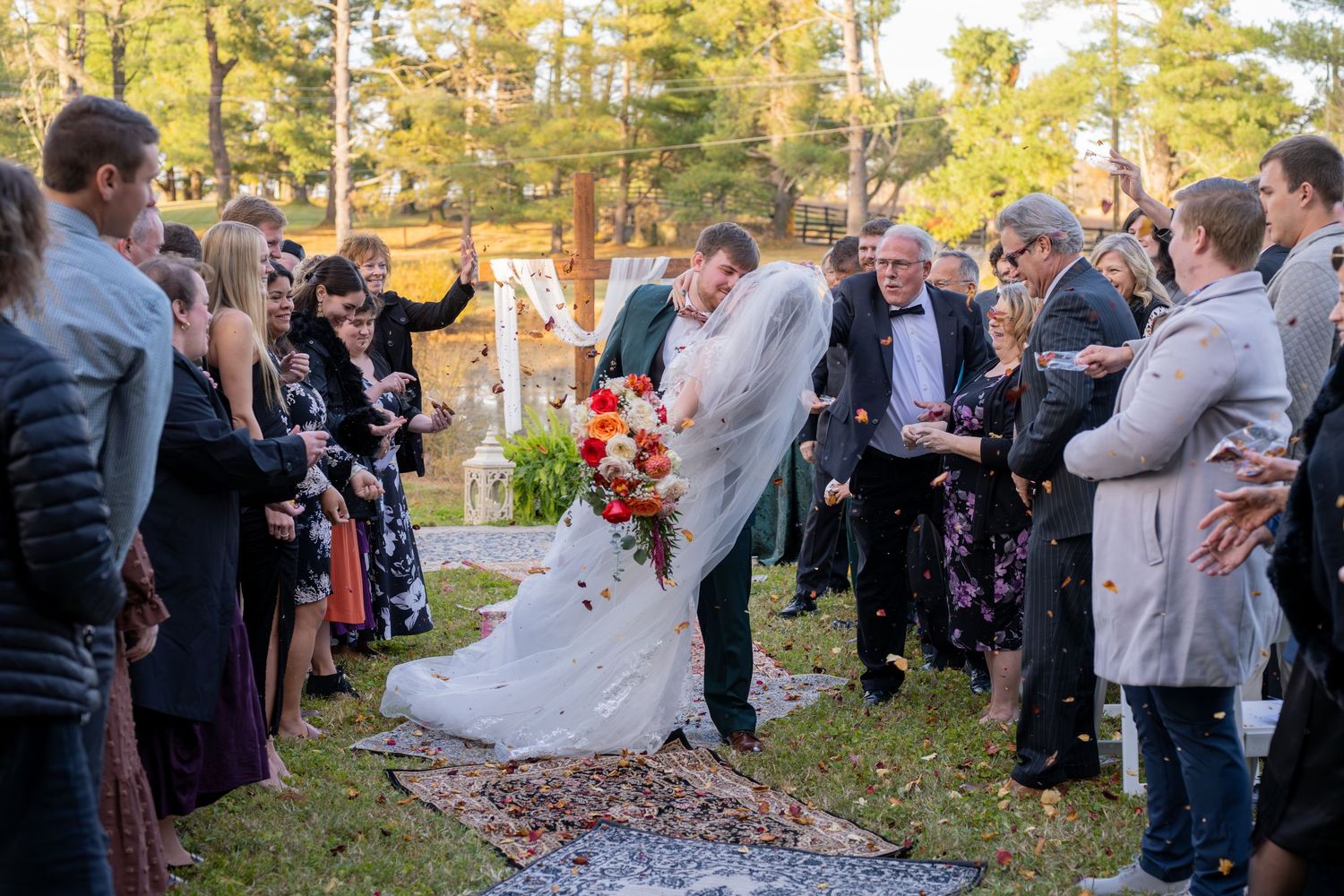 A couple shares a romantic kiss during their outdoor autumn wedding ceremony as guests look on in a wooded setting.