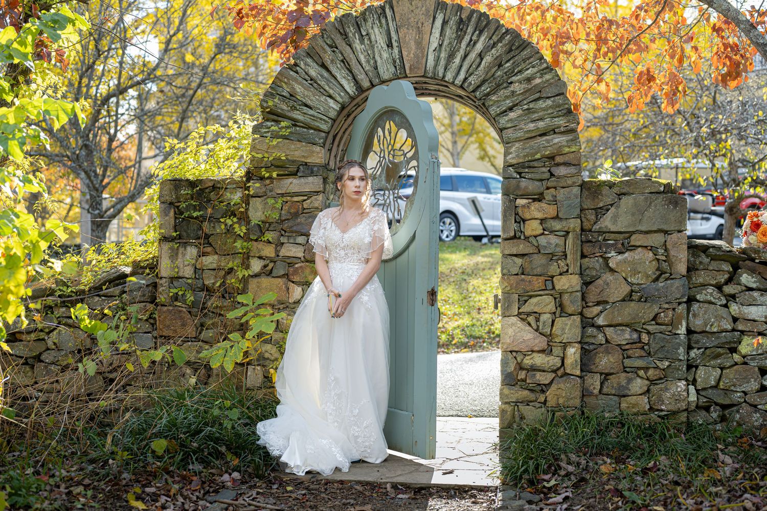 A bride in a white wedding dress poses against a rustic stone wall with autumn foliage in the background.