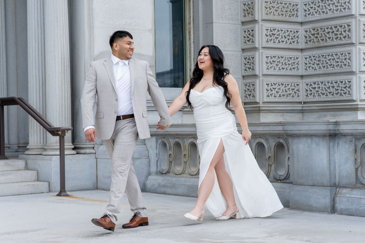 Couple in wedding attire holding hands walking outside a grand stone building, bride in white gown, groom in tan suit.