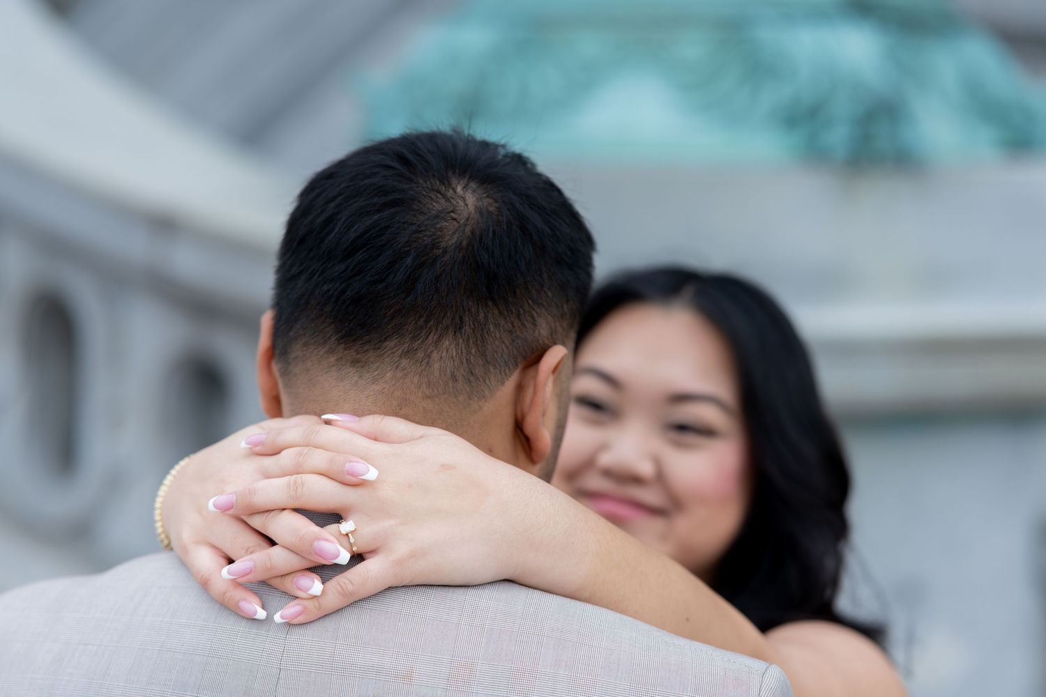 A woman smiles while embracing a man, showing off her engagement ring, with a blurred architectural background.