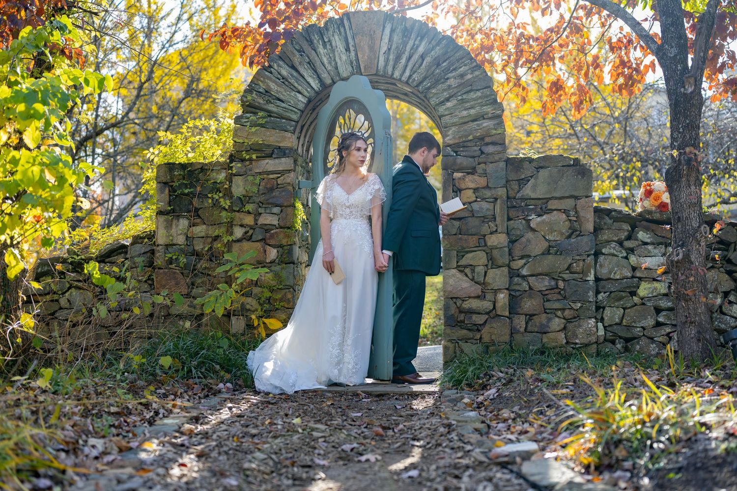 A couple stands together in a picturesque autumn garden setting with stone walls and colorful fall foliage.