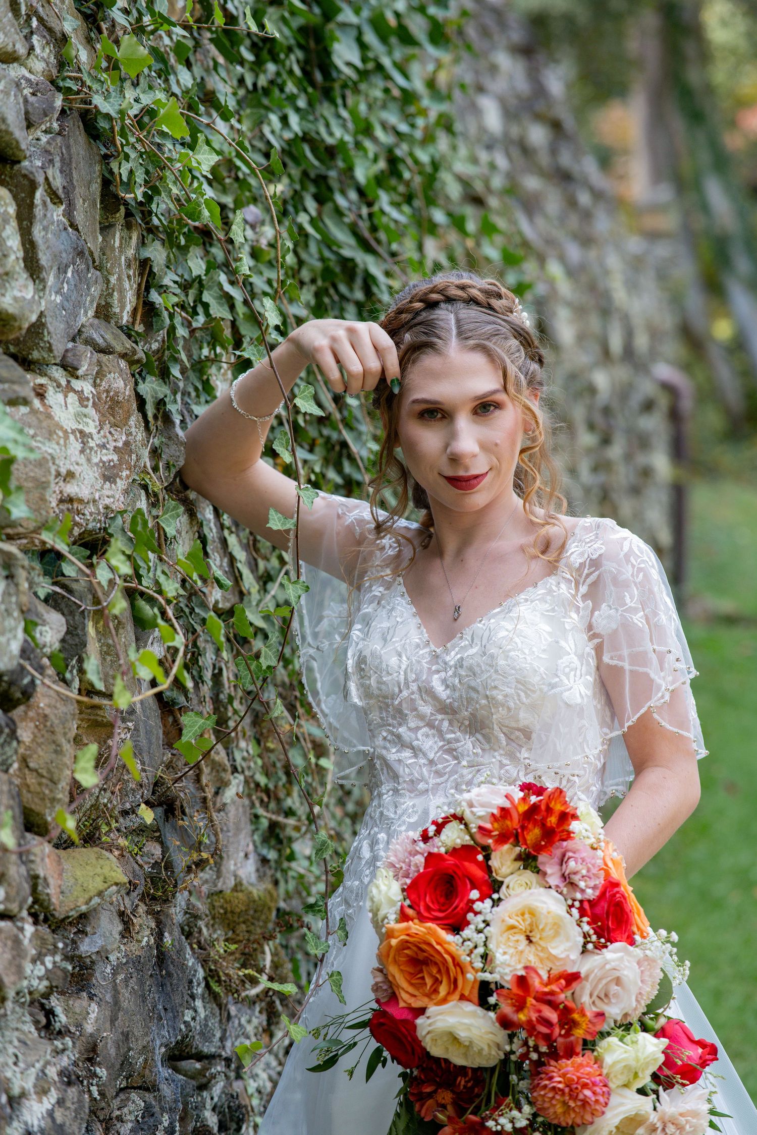 A bride in a lace wedding dress poses against an ivy-covered stone wall with a vibrant red and orange bouquet.