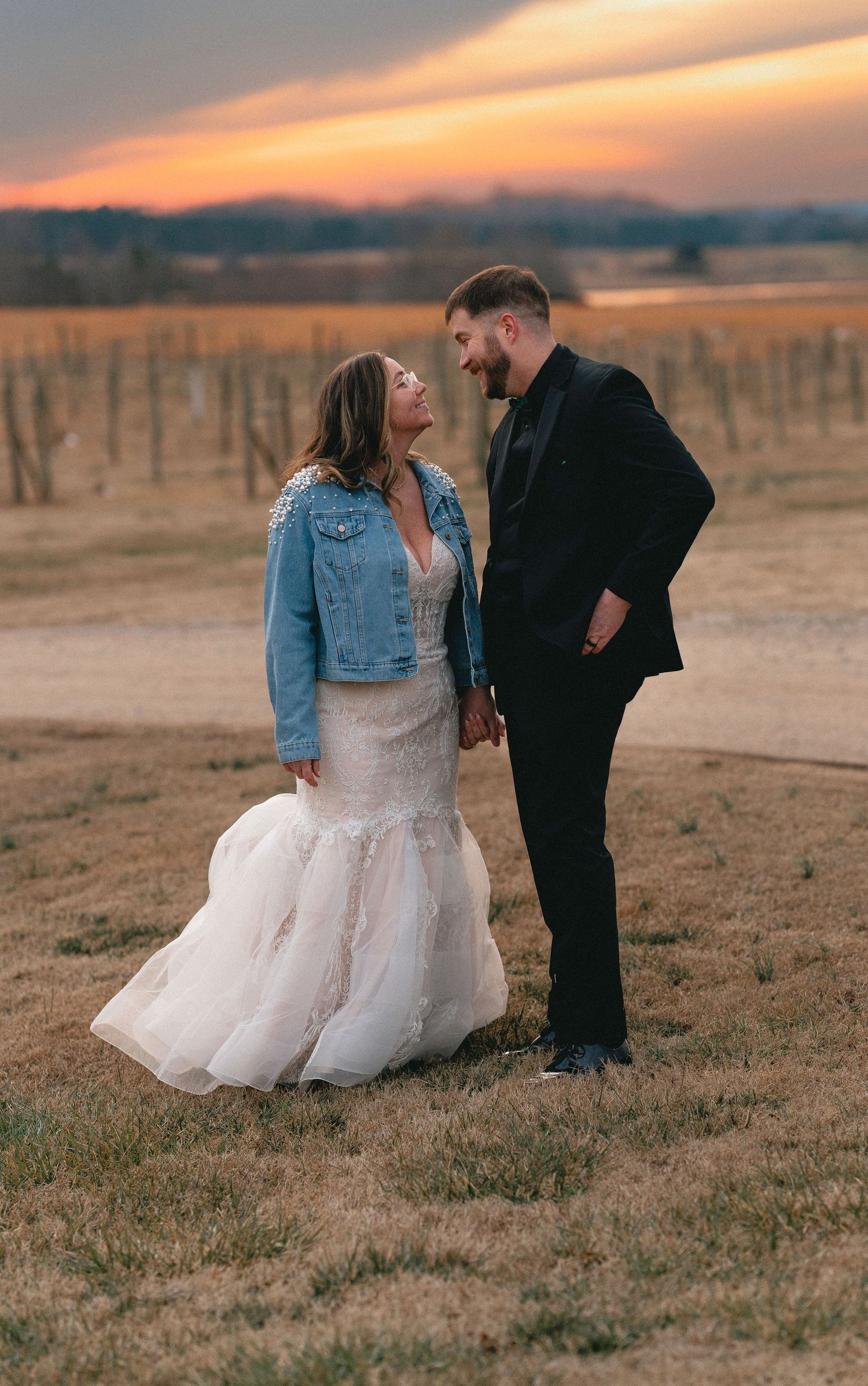 Couple takes romantic sunset photos in a field wearing wedding attire and denim jacket.