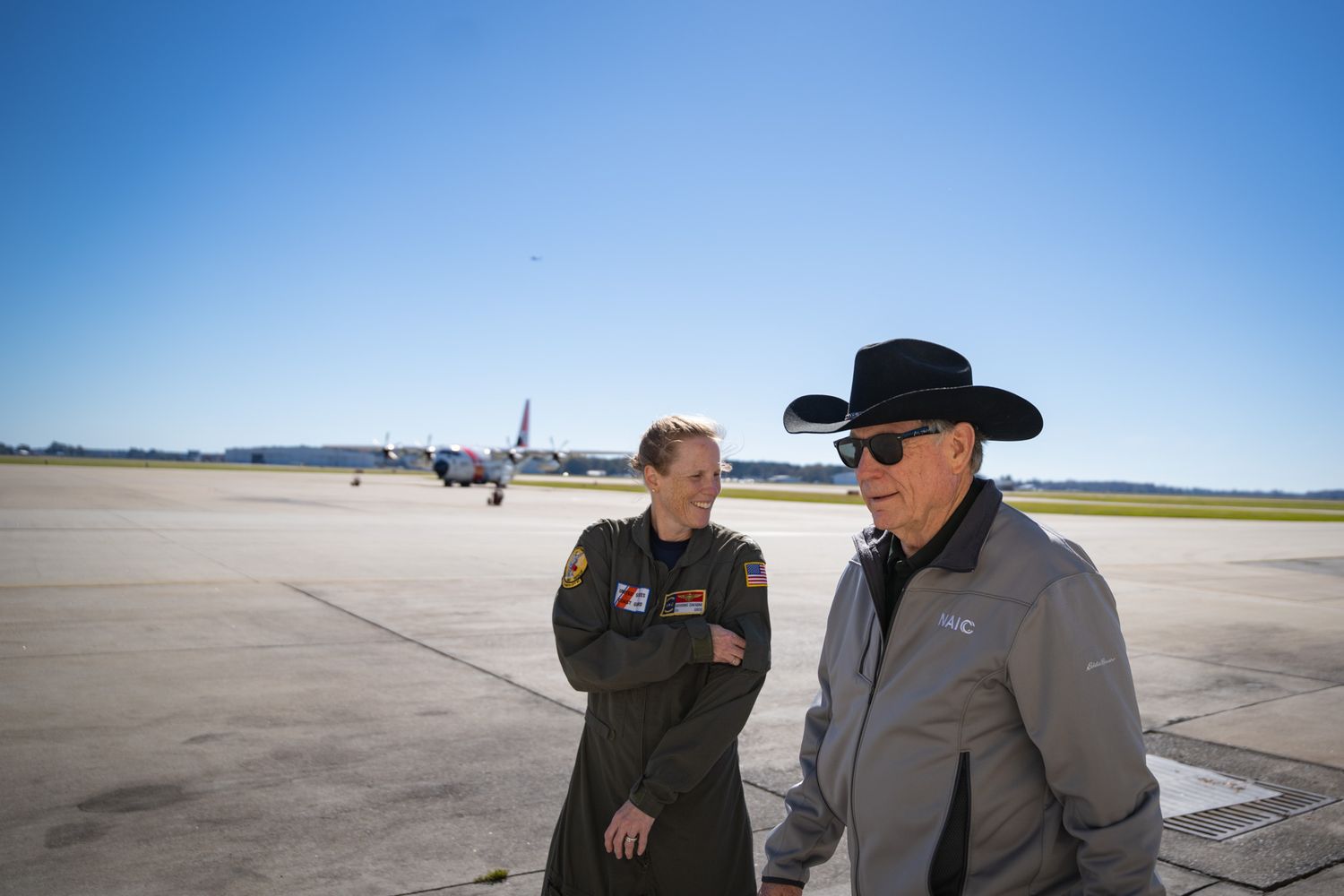 People on an airport tarmac with aircraft visible in the bright sunlight background.