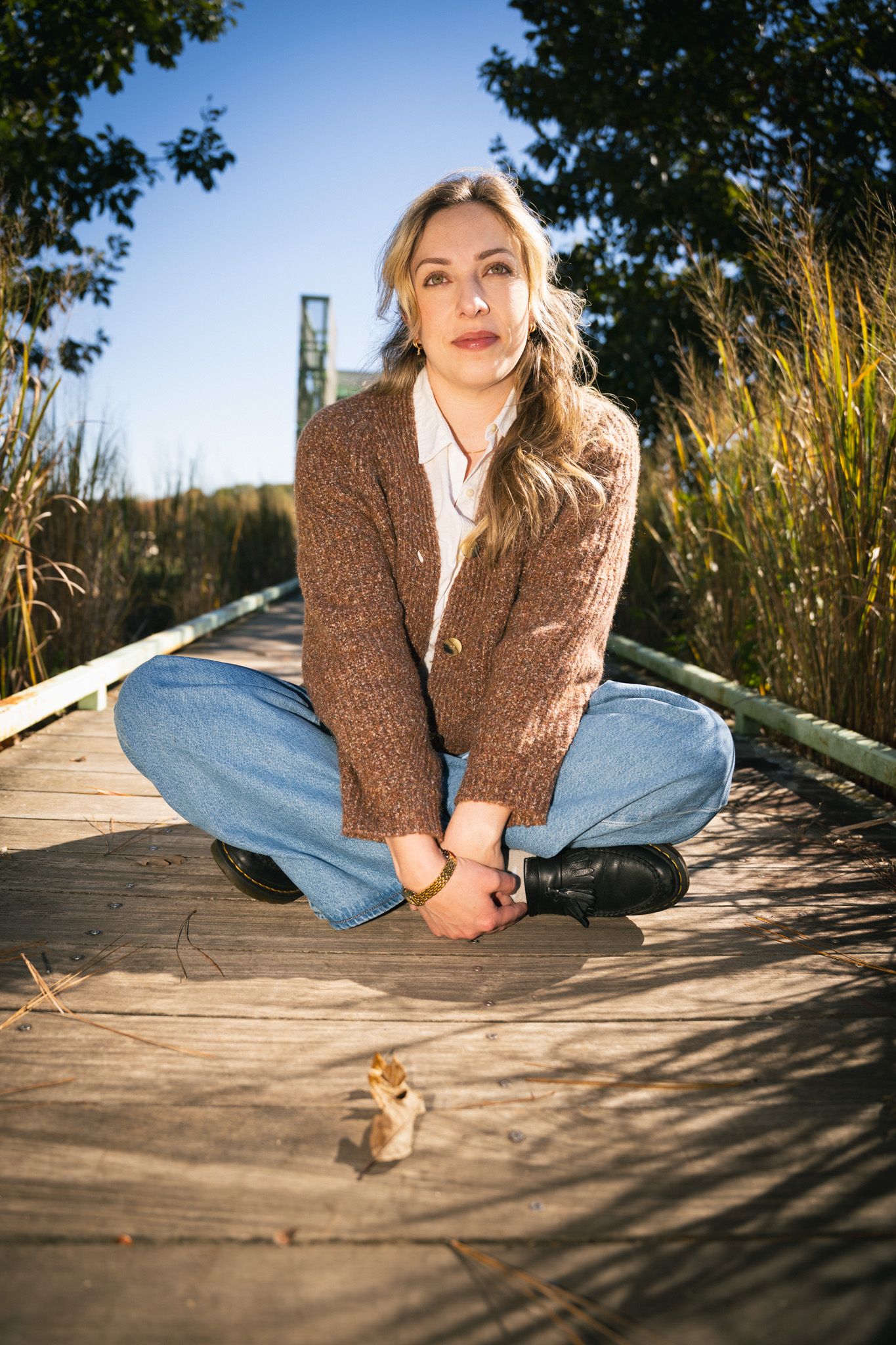 Person in brown sweater and jeans sitting on wooden boardwalk during golden hour.