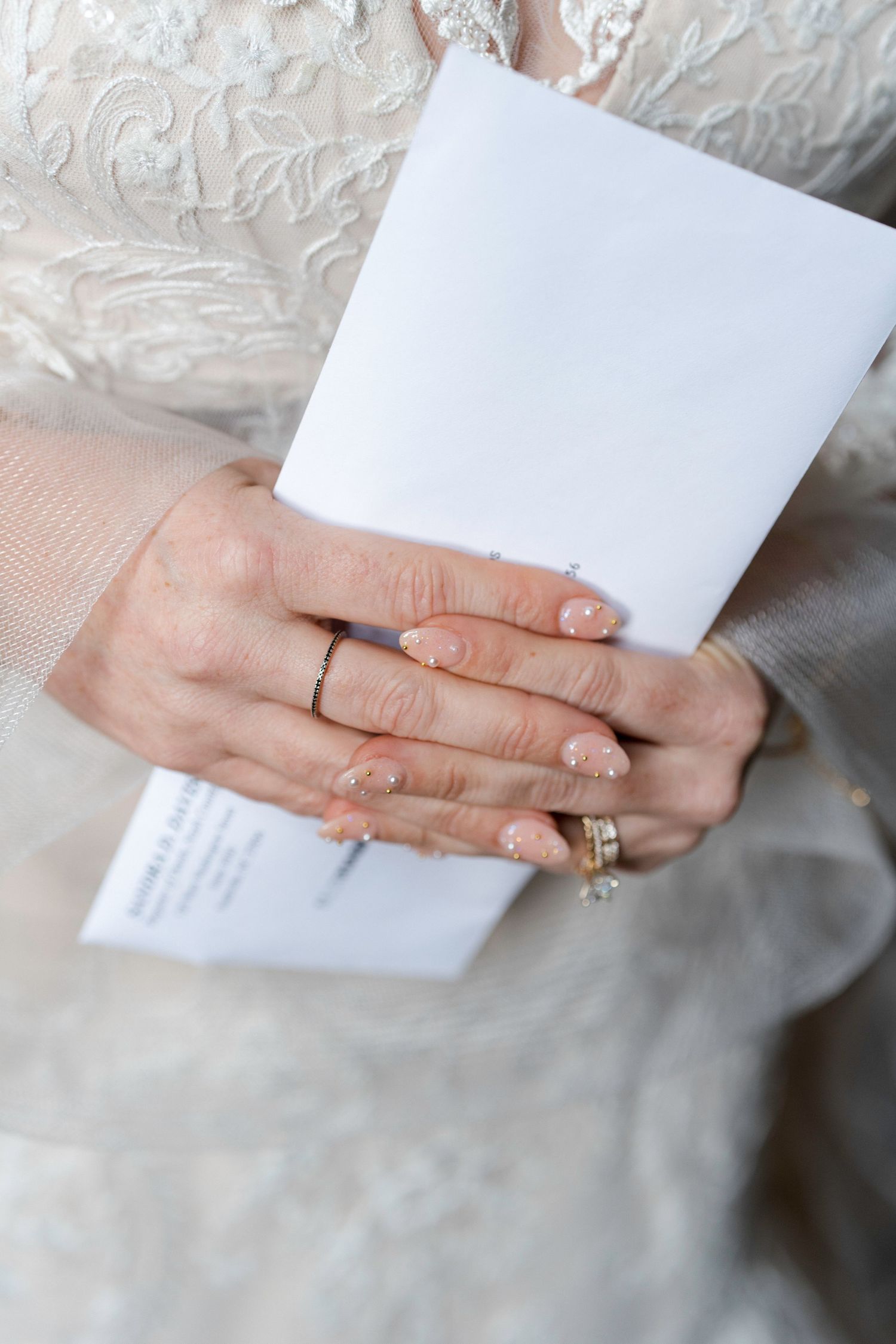 Close-up of hands holding white paper with wedding ring visible against lace dress fabric.