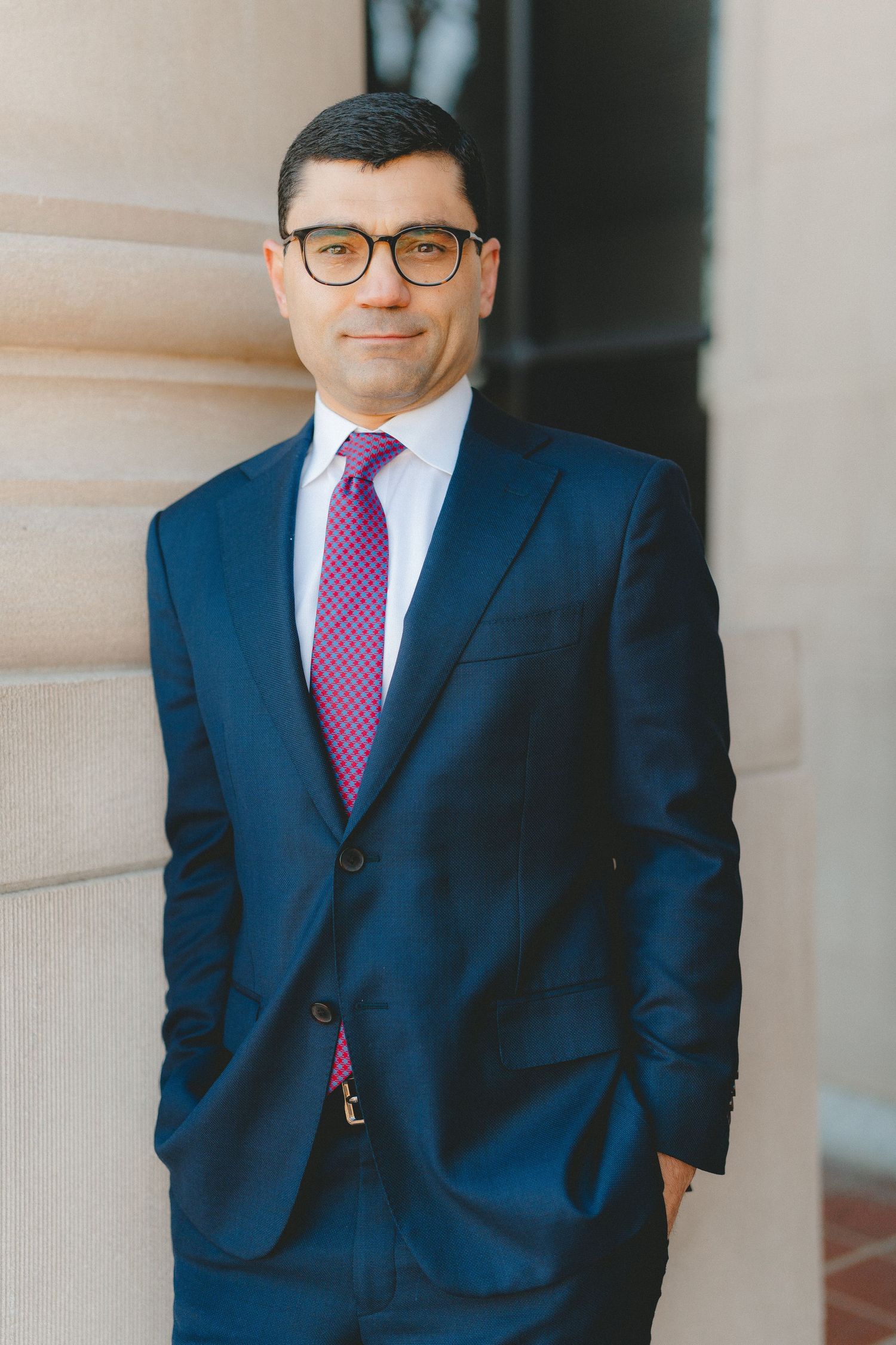 Professional headshot of person in navy suit with purple tie against light background.