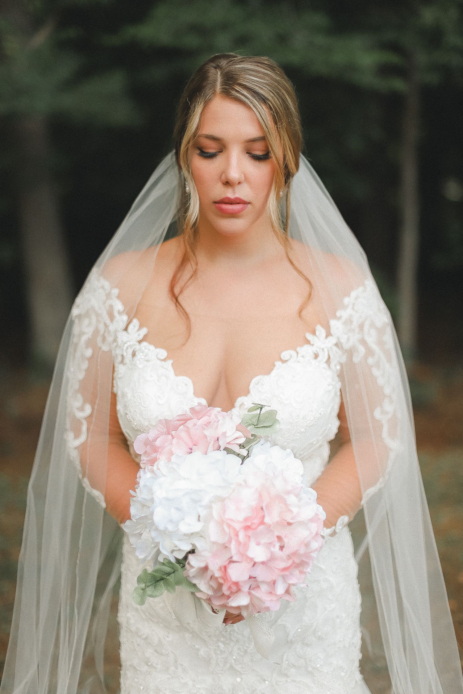 Bride in detailed lace wedding gown holds pink and white bouquet in outdoor bridal portraits.