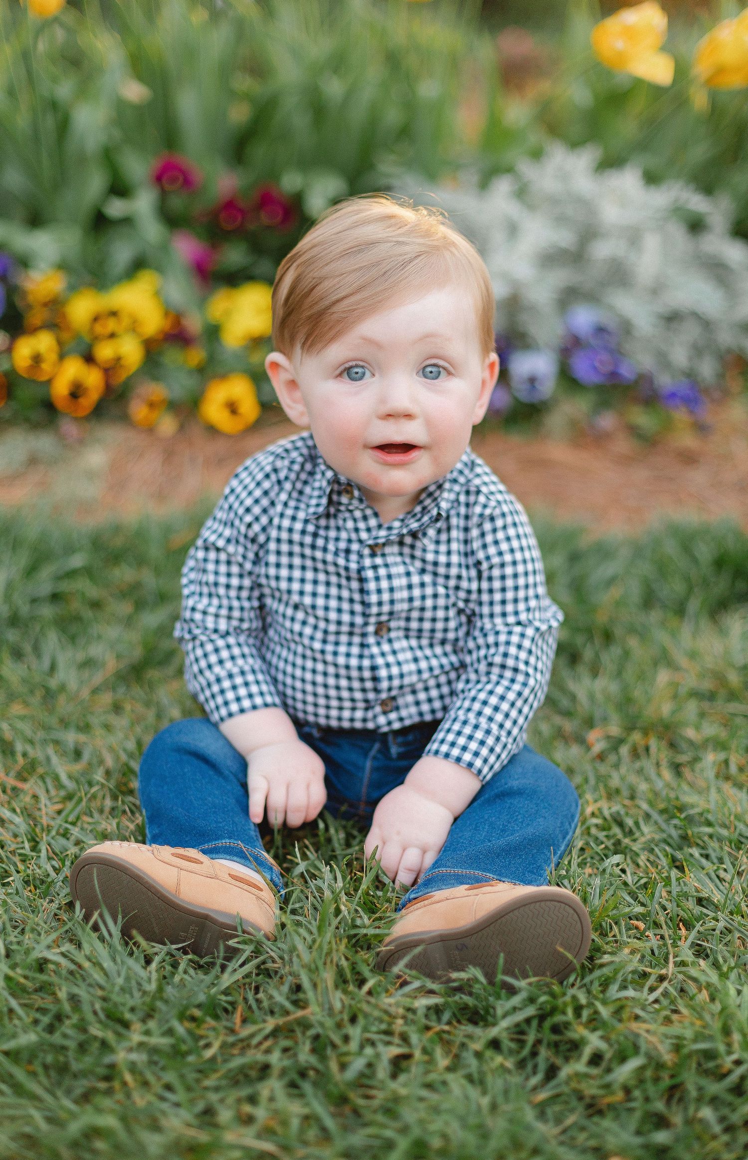 Toddler in checkered shirt and jeans sits on grass surrounded by colorful flowers.