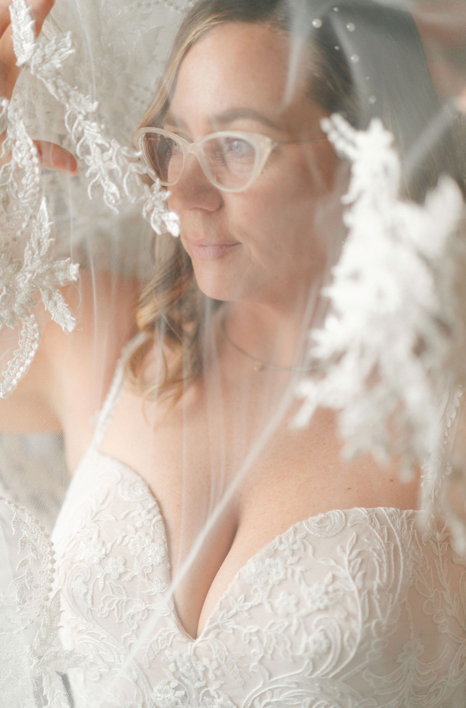 Ethereal bridal portrait through delicate lace veil showing cat-eye glasses and white wedding dress details.