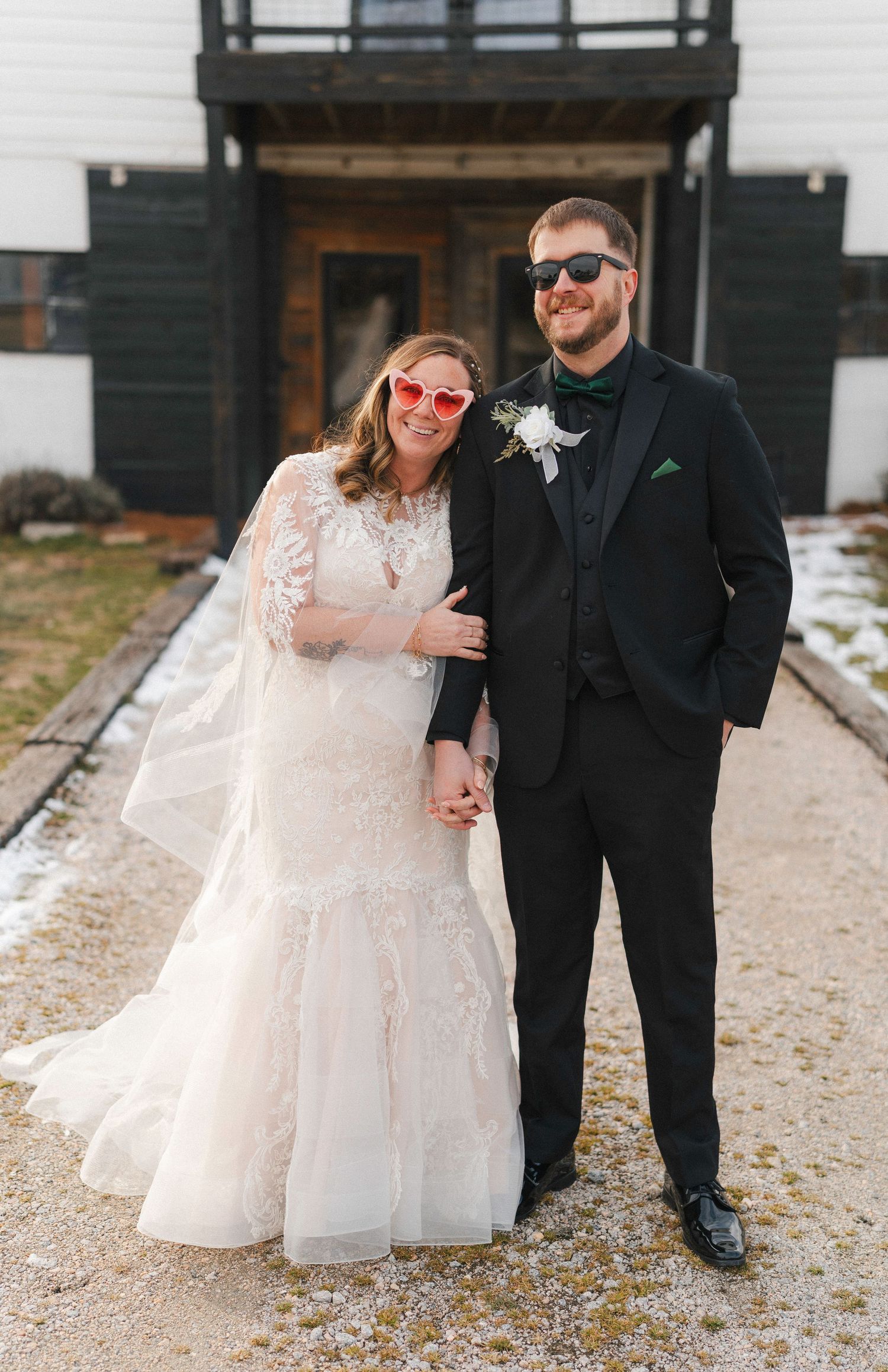 Couple poses for winter wedding photos on a gravel path outside a modern building.