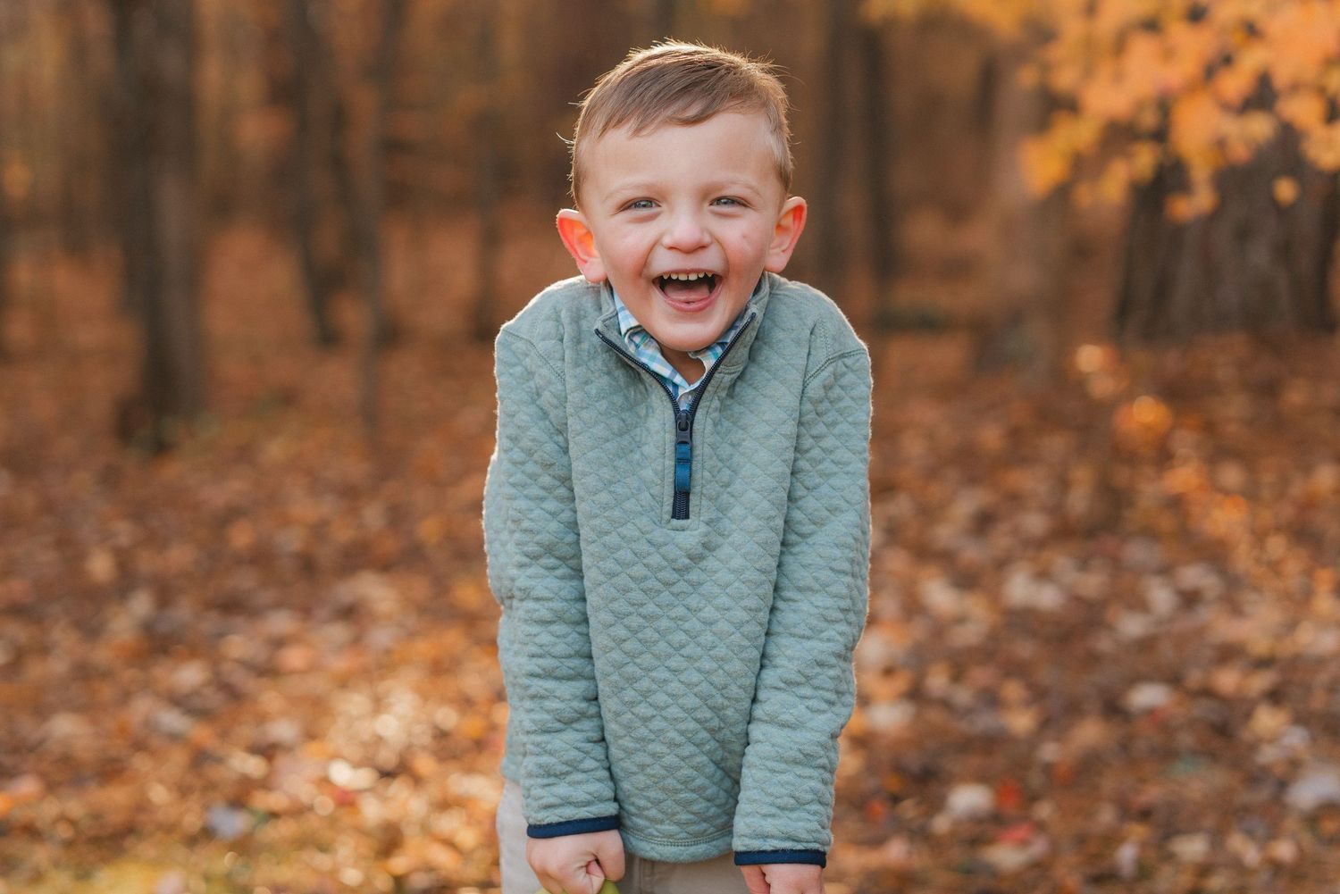 Child playing and laughing in fallen autumn leaves wearing a teal quilted sweater.
