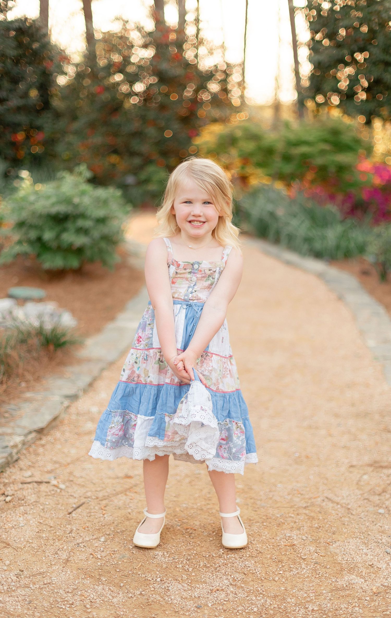 Young child in blue and floral tiered dress twirls on garden path at sunset.