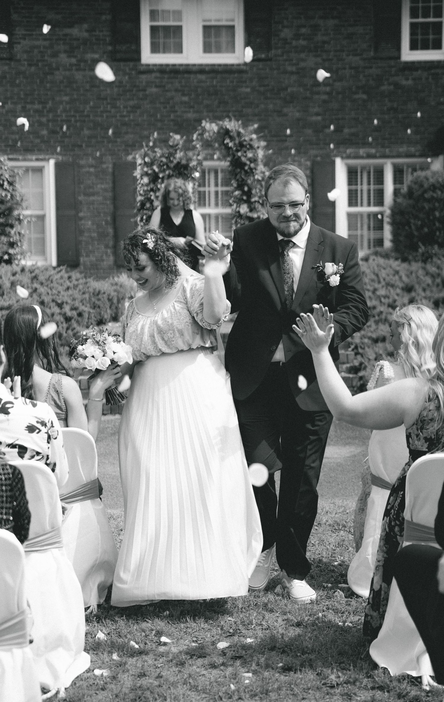 Outdoor wedding celebration with guests in white chairs against a brick building backdrop.
