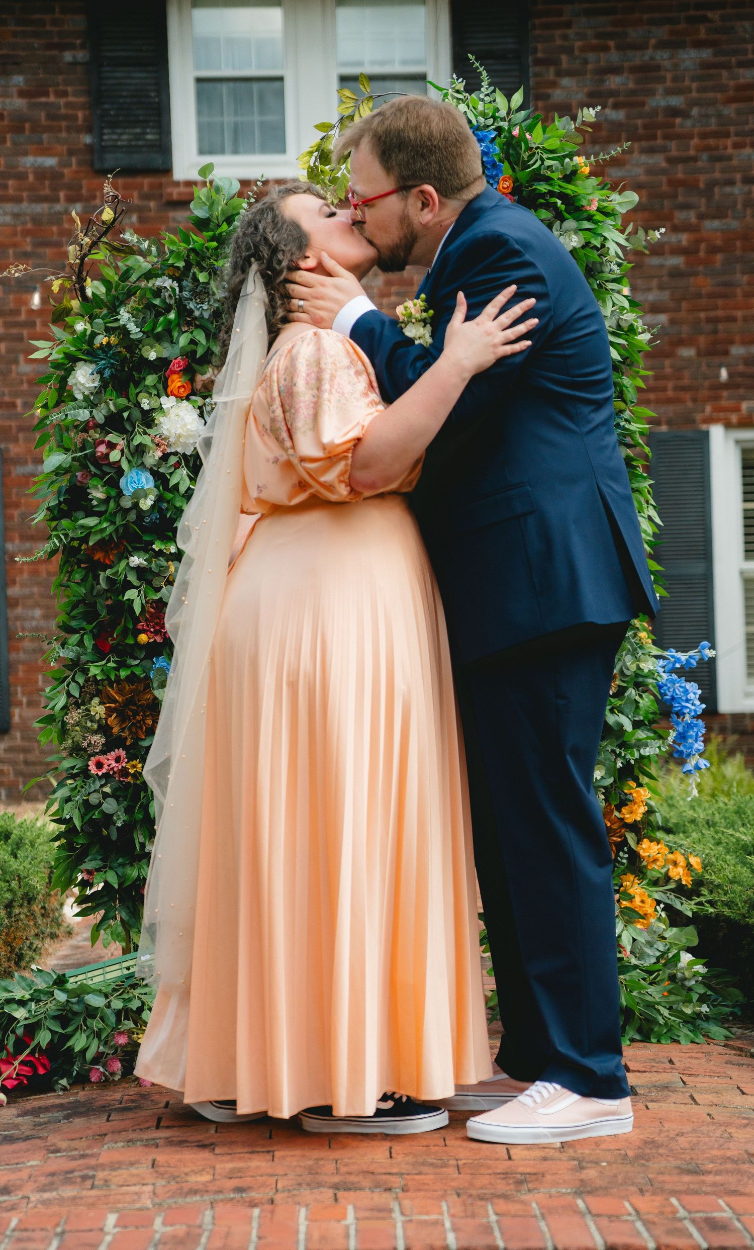Wedding couple shares romantic kiss under floral arch against brick building.