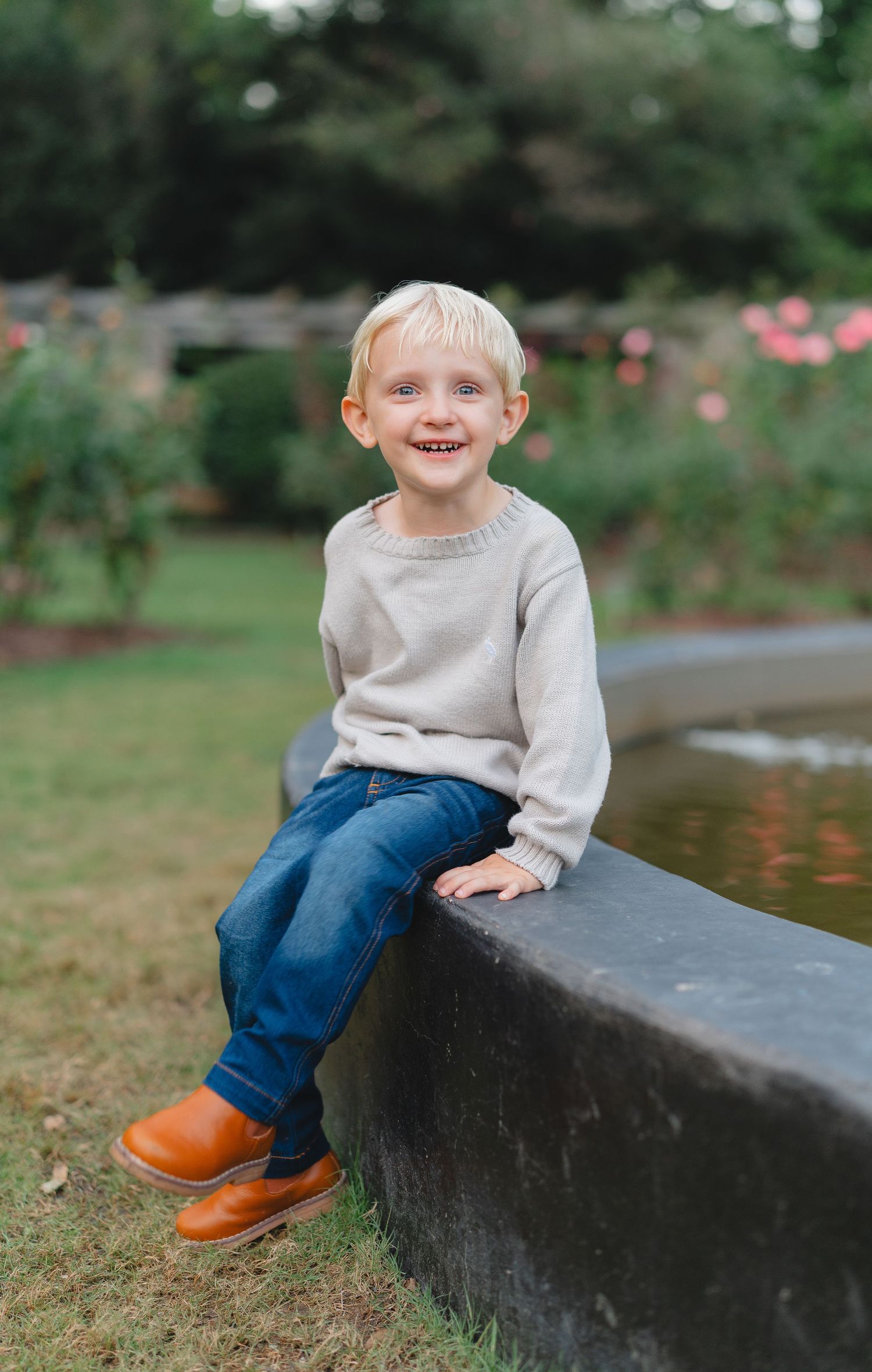 Young child in gray sweater and jeans sits smiling on stone wall in garden setting.