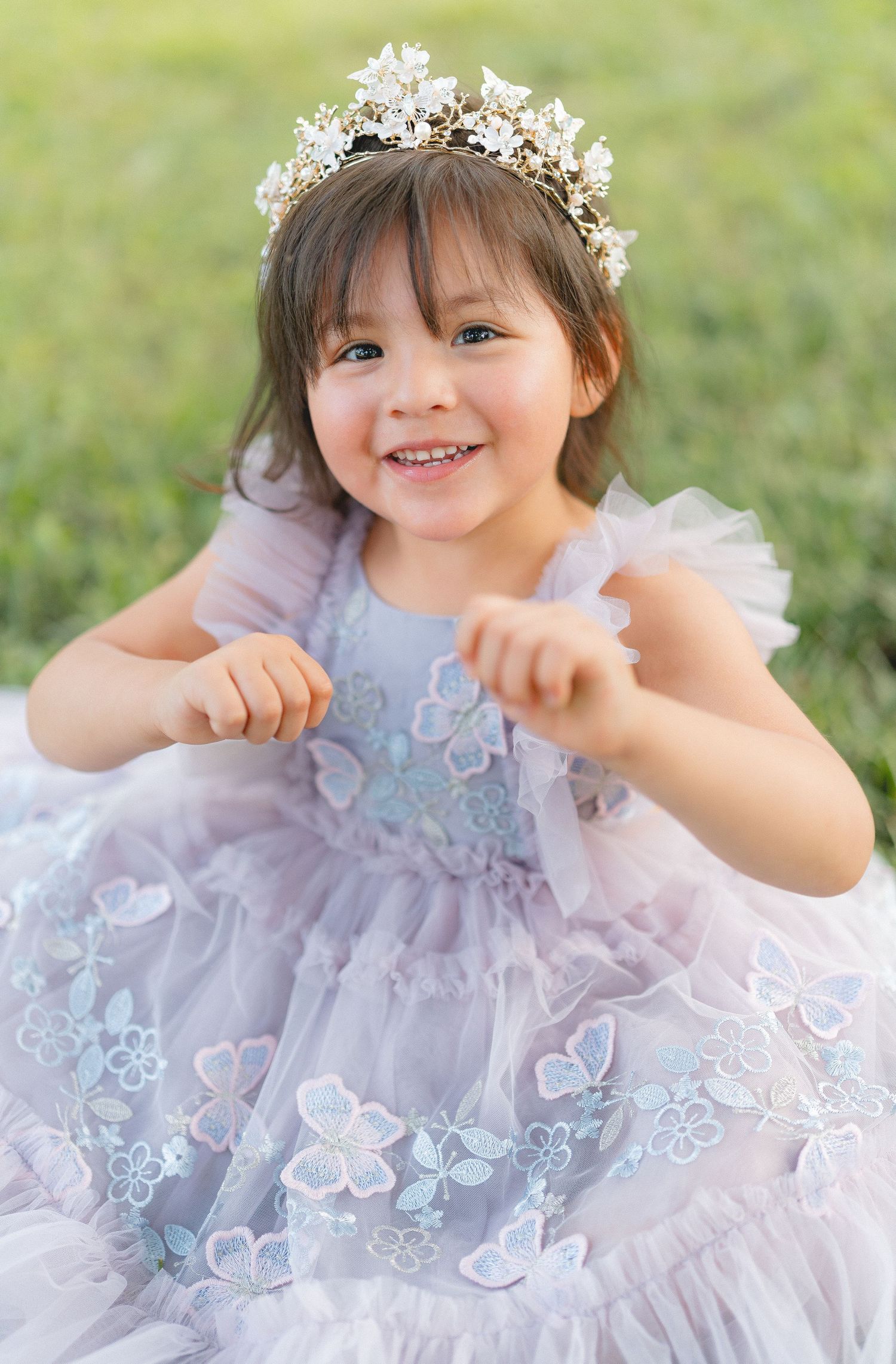 A child in a dreamy lavender tulle dress with butterfly appliques poses joyfully in a grassy outdoor setting.