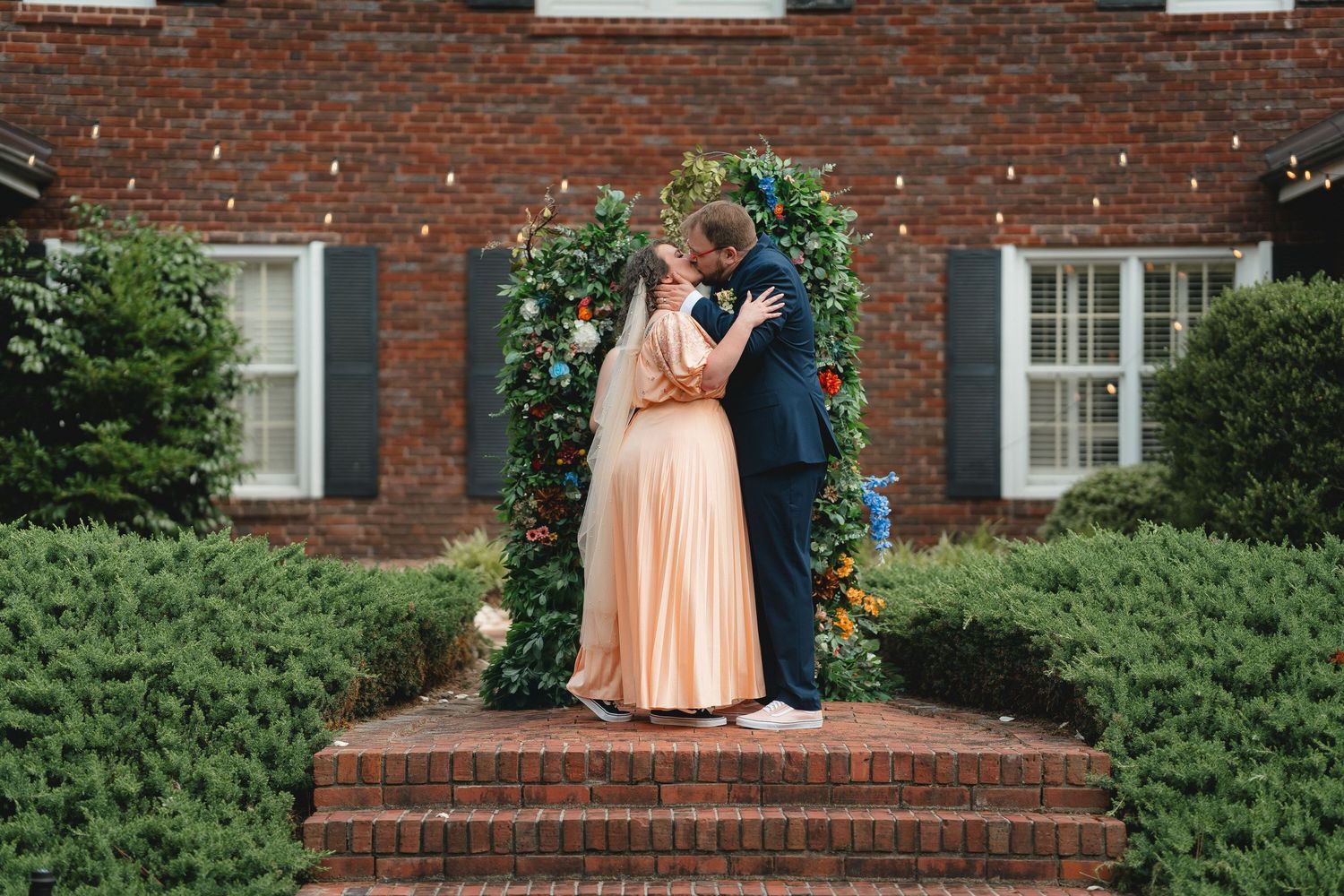 Couple posing on brick steps outside traditional brick building with greenery.