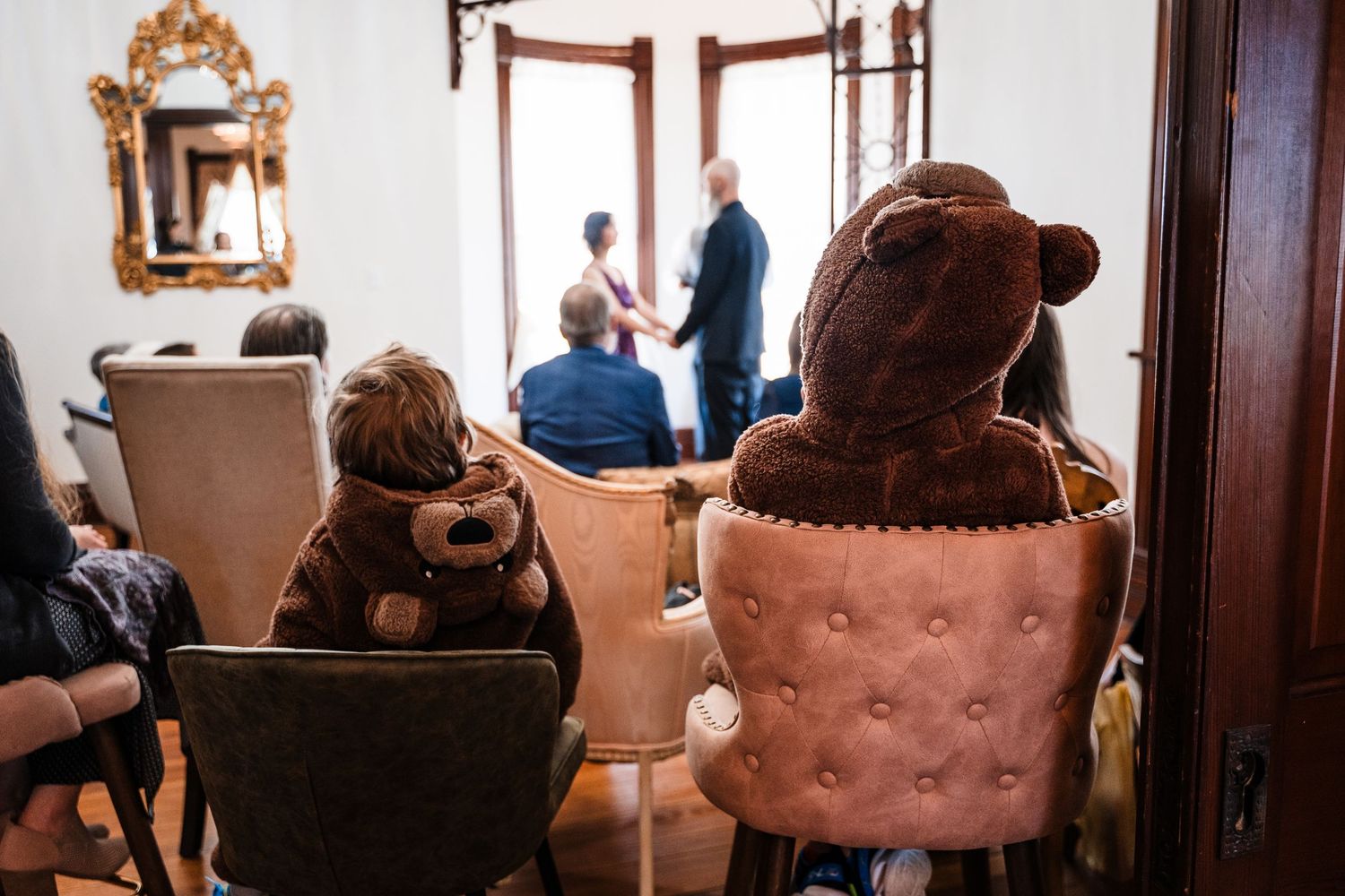 Child and teddy bear seated in chairs watching a couple standing by a doorway in an elegant room.