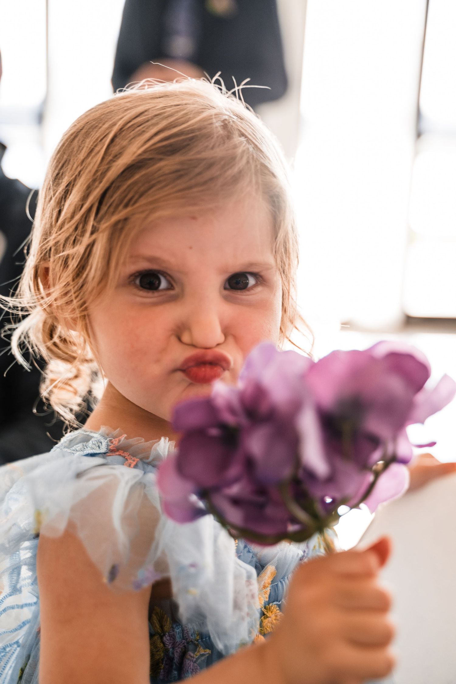 Adorable young flower girl in blue dress making kissy face while holding purple flowers at wedding.