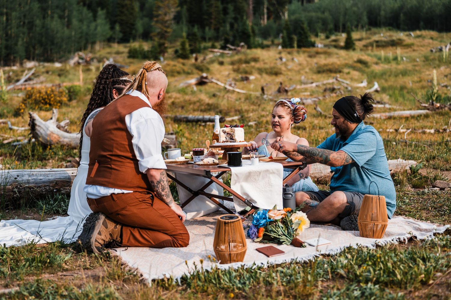 A bohemian outdoor elopement picnic with four people gathered around a rustic table in a mountain meadow.