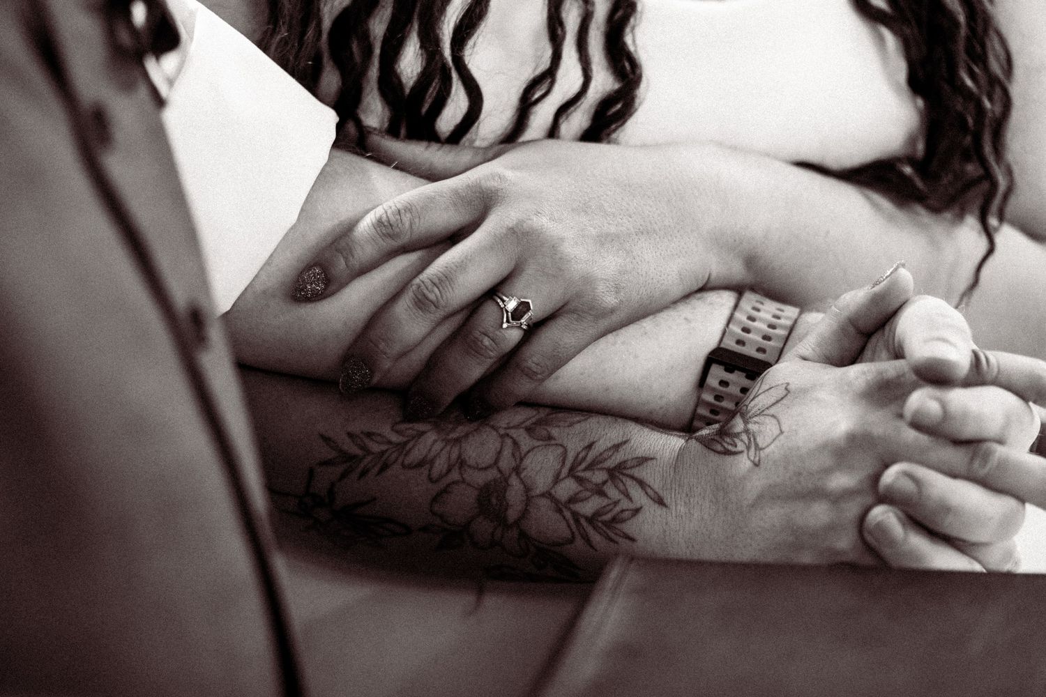Close-up black and white photo of couple holding hands showing wedding rings and floral tattoos.