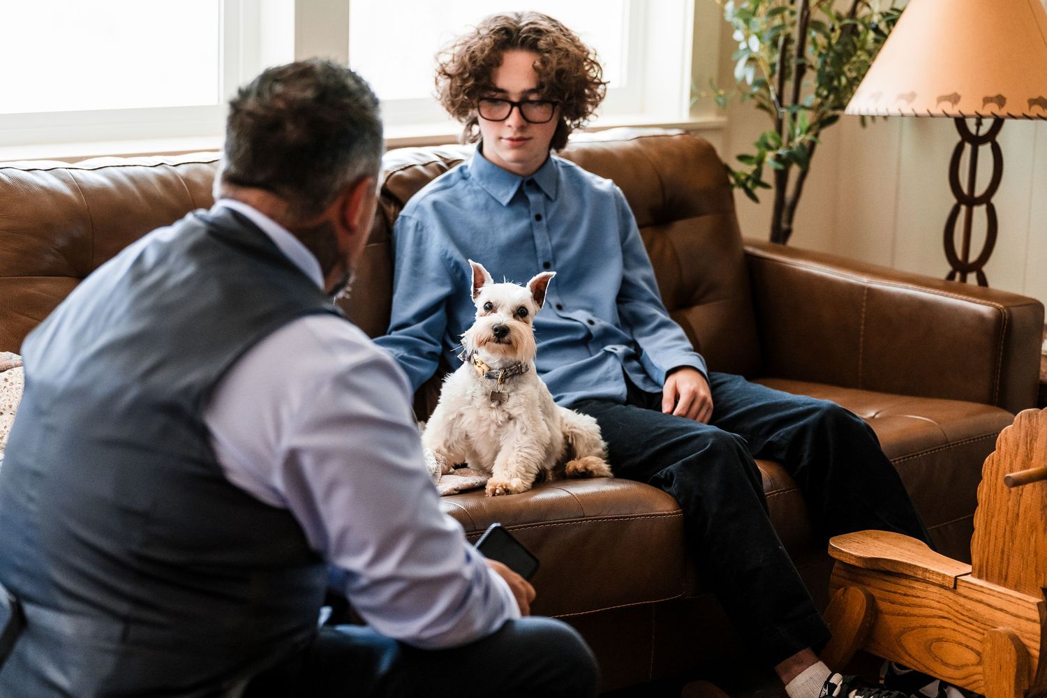 Young person with curly hair and glasses sits on leather sofa with small white terrier dog during therapy session.
