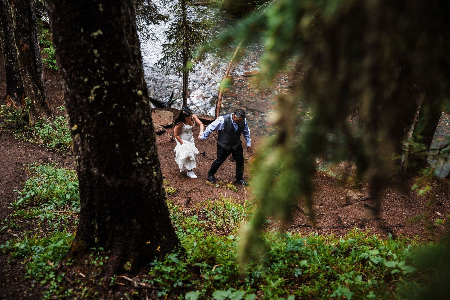 Bride and groom walk hand in hand through lush green forest during outdoor wedding.