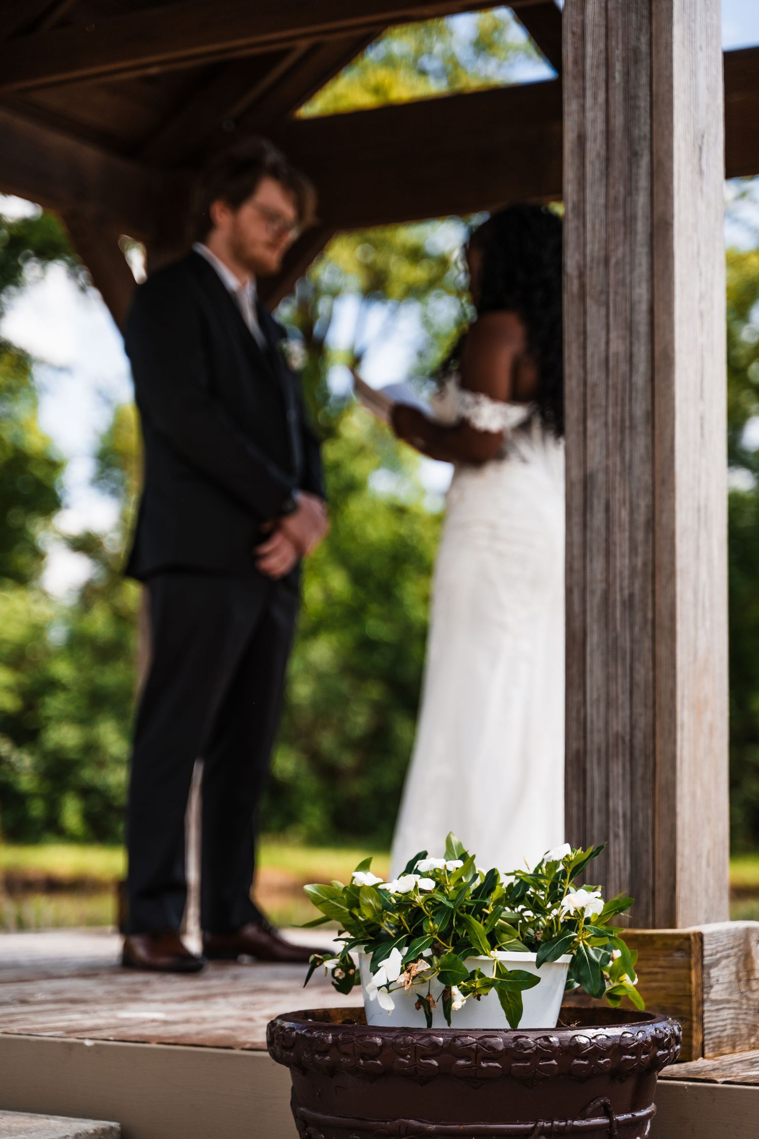 Bride and groom sharing intimate moment under wooden gazebo with white flowers in foreground.
