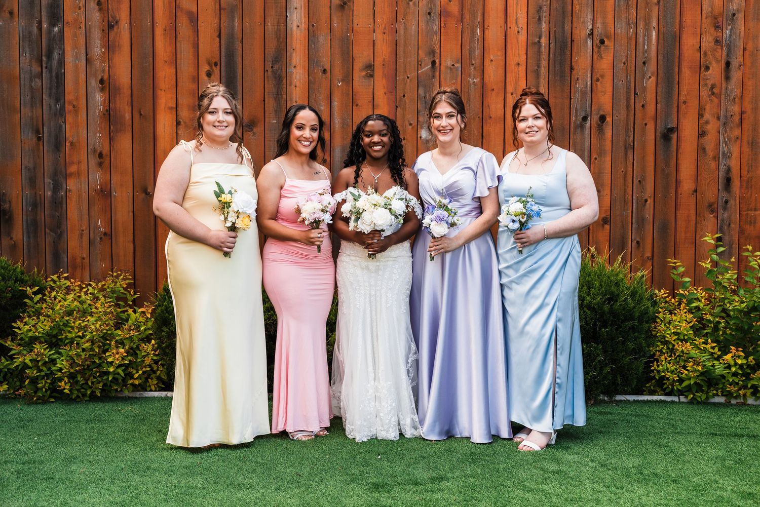 Bride with four bridesmaids in colorful mismatched gowns posing against a wooden fence backdrop.