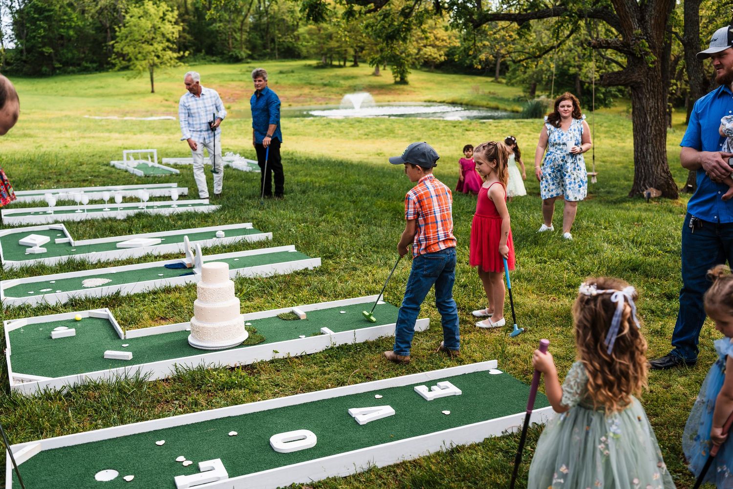 Children and guests enjoy outdoor mini golf wedding lawn game with white wedding cake as a hole marker.