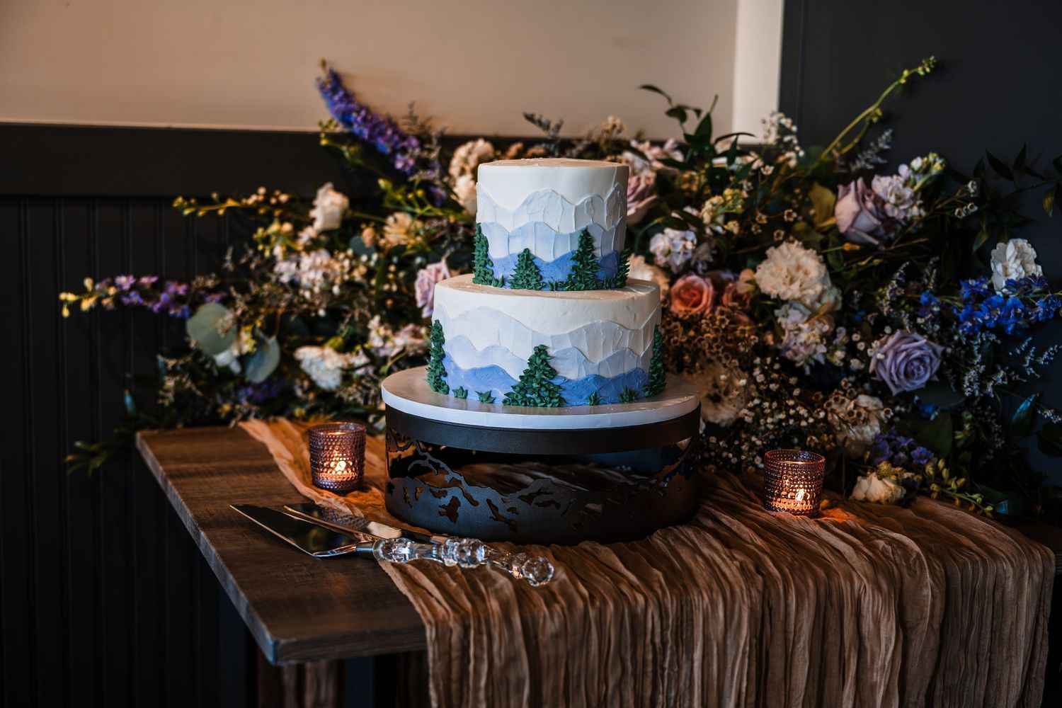 Three-tier mountain landscape wedding cake surrounded by colorful floral arrangements on rustic table.