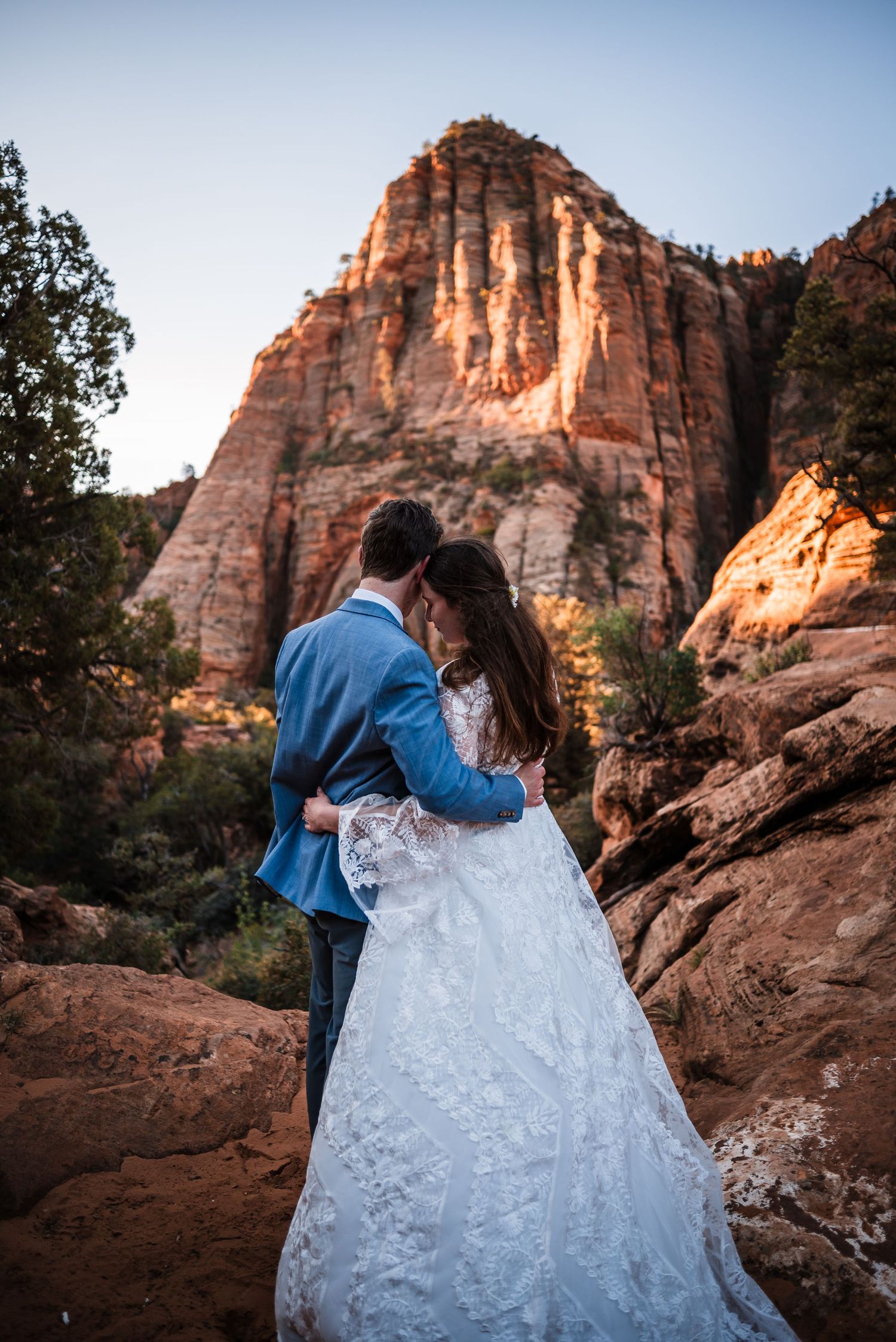 Bride and groom embracing at sunset with dramatic Zion National Park red rock formations in background.