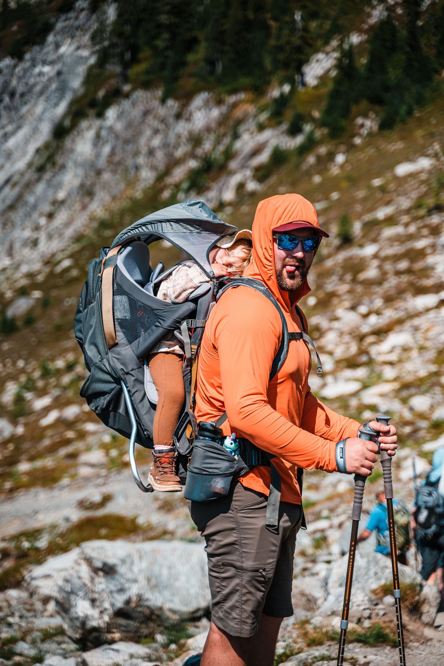Hiker in bright orange jacket traverses snowy mountain terrain with hiking poles.