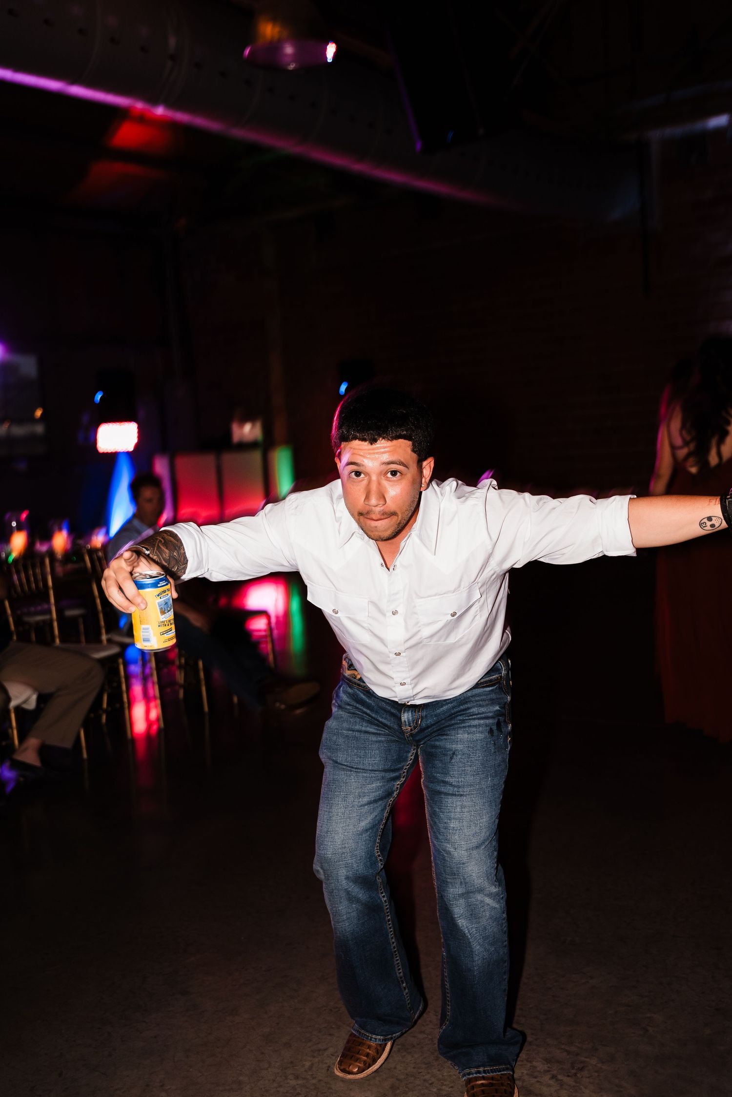 Person dancing at reception in white shirt and jeans with colorful lights in background.