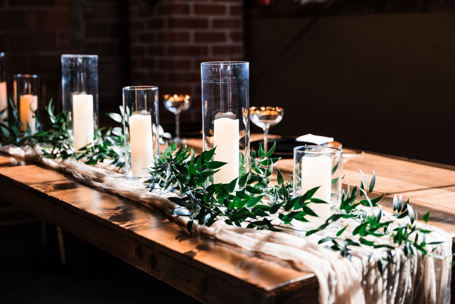 An elegant table setting with glass cylinders holding candles and green foliage runners.