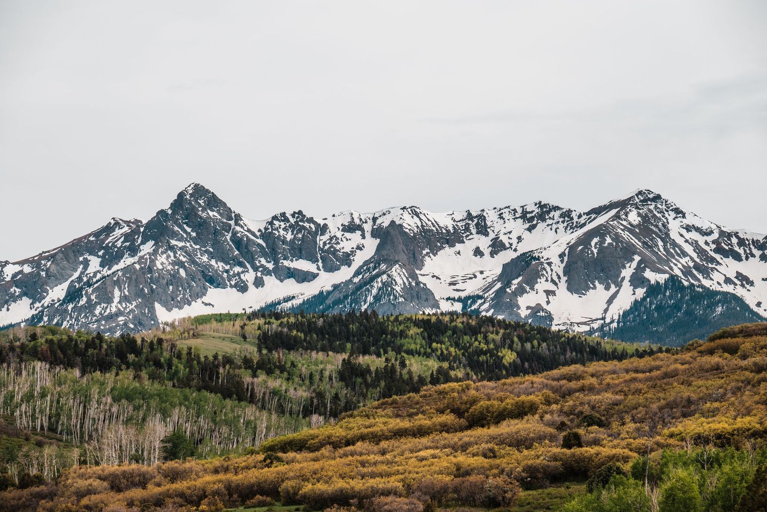 Snow-capped mountain peaks rise above autumn colored slopes in a rugged mountain landscape.