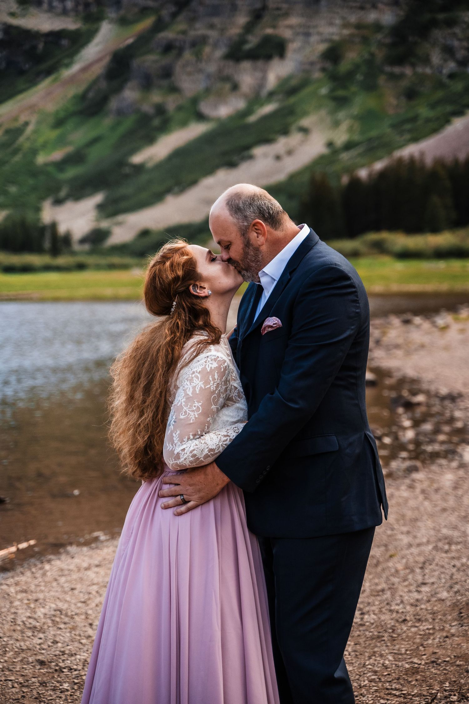 Romantic kiss between newlyweds beside a mountain lake with dramatic peaks in the background.