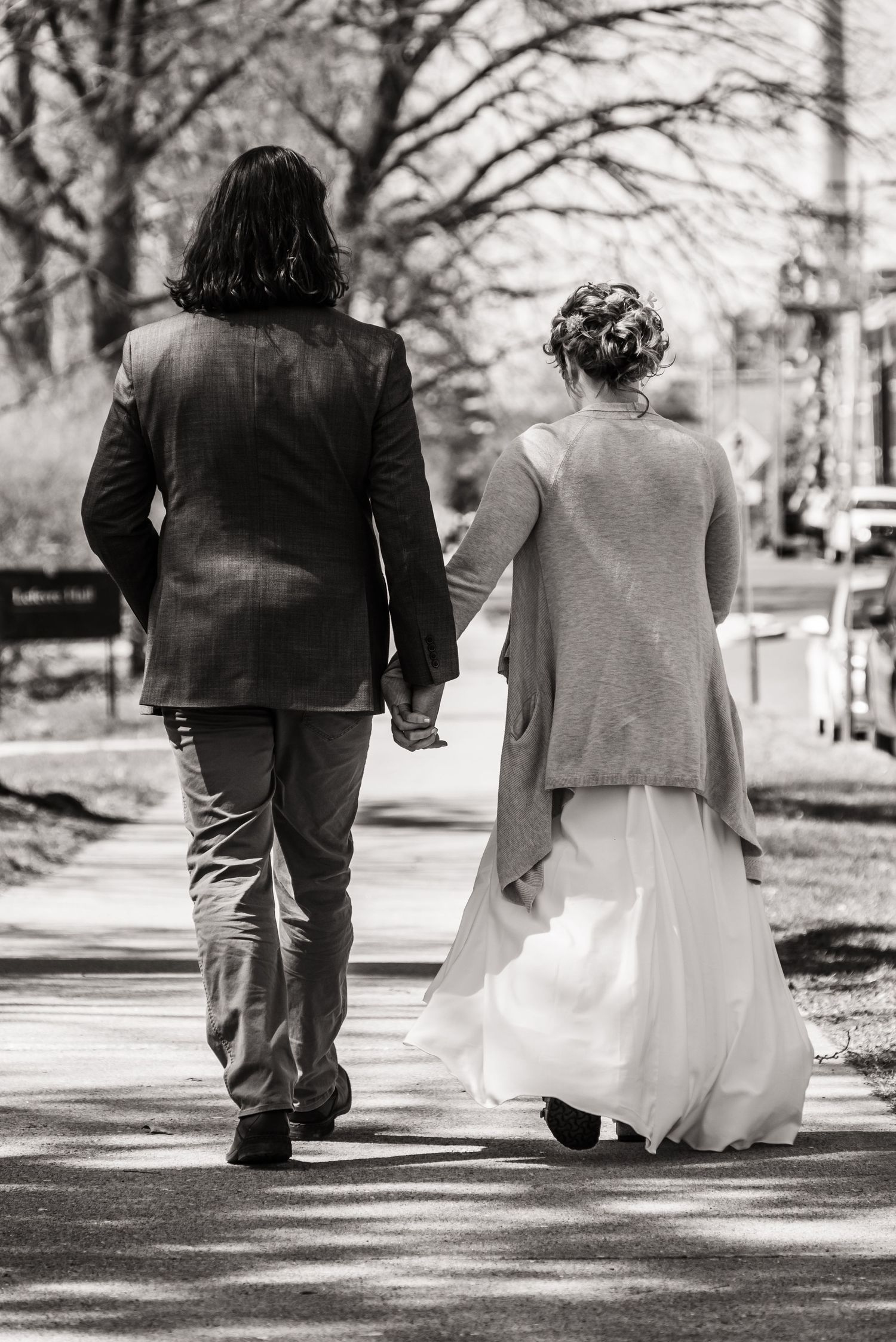 Black and white photo of couple walking hand in hand down tree-lined path.