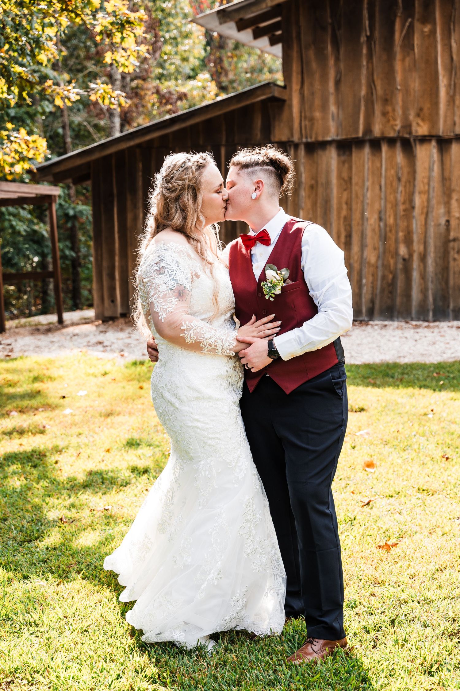 Wedding couple embraces in front of rustic barn on sunny day.