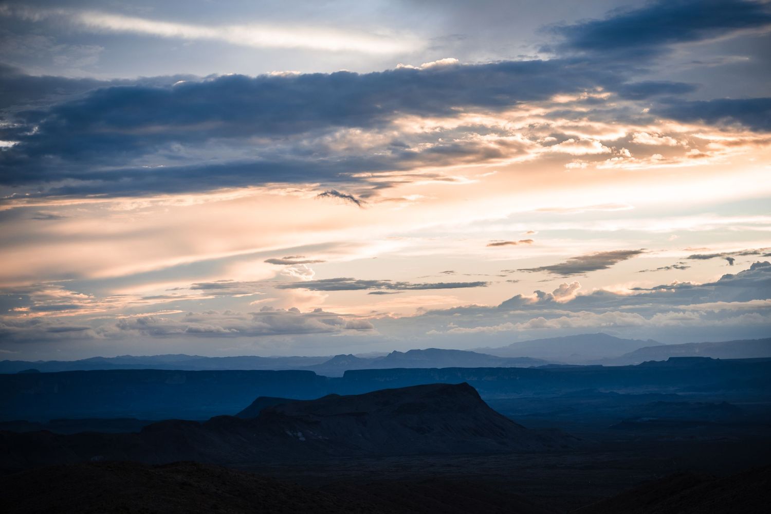 Silhouetted mesa landscape at sunset with layered mountains and dramatic cloudy sky.