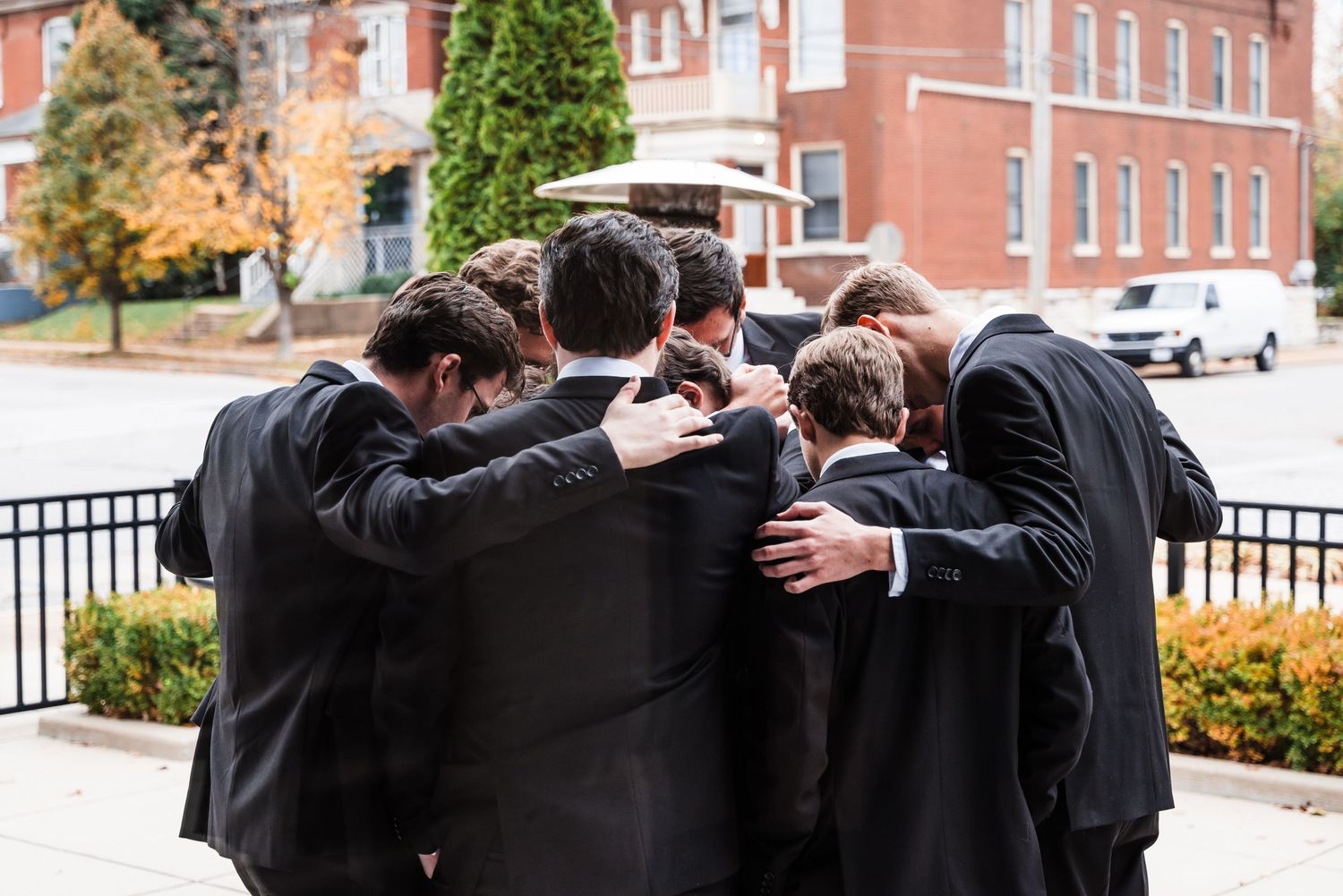 Groomsmen huddle together in a circle outside a brick building.