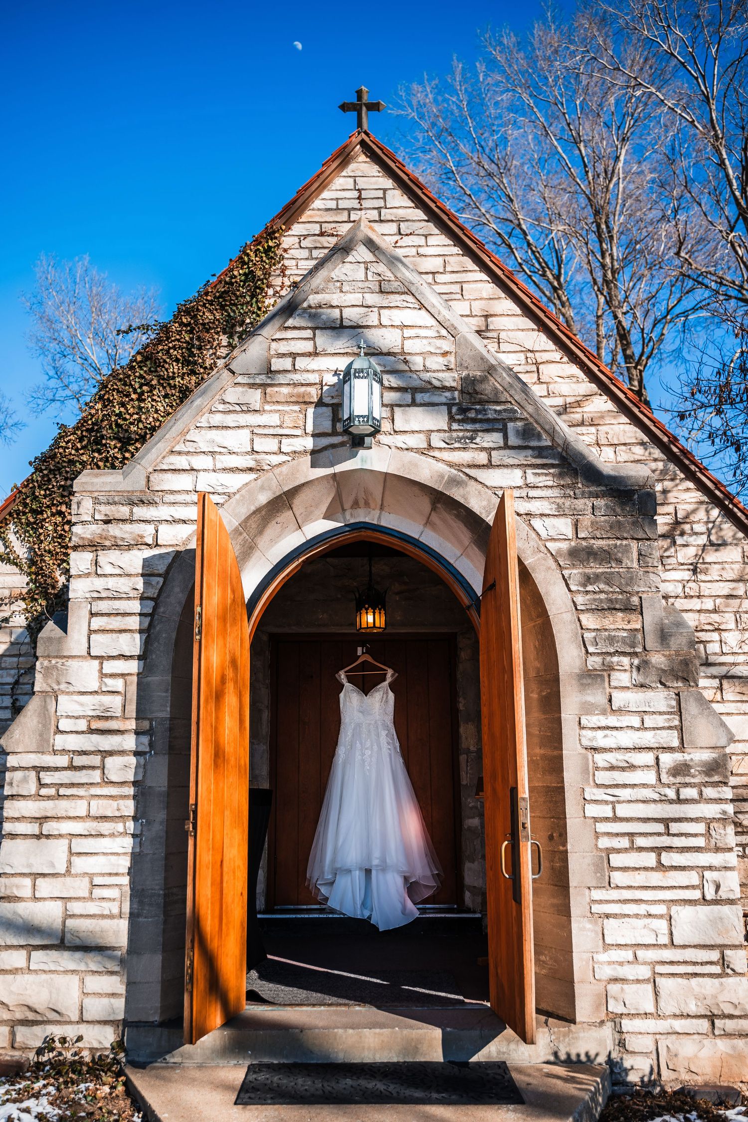 A white wedding dress hangs in the doorway of a stone chapel with wooden doors and a cross on top.