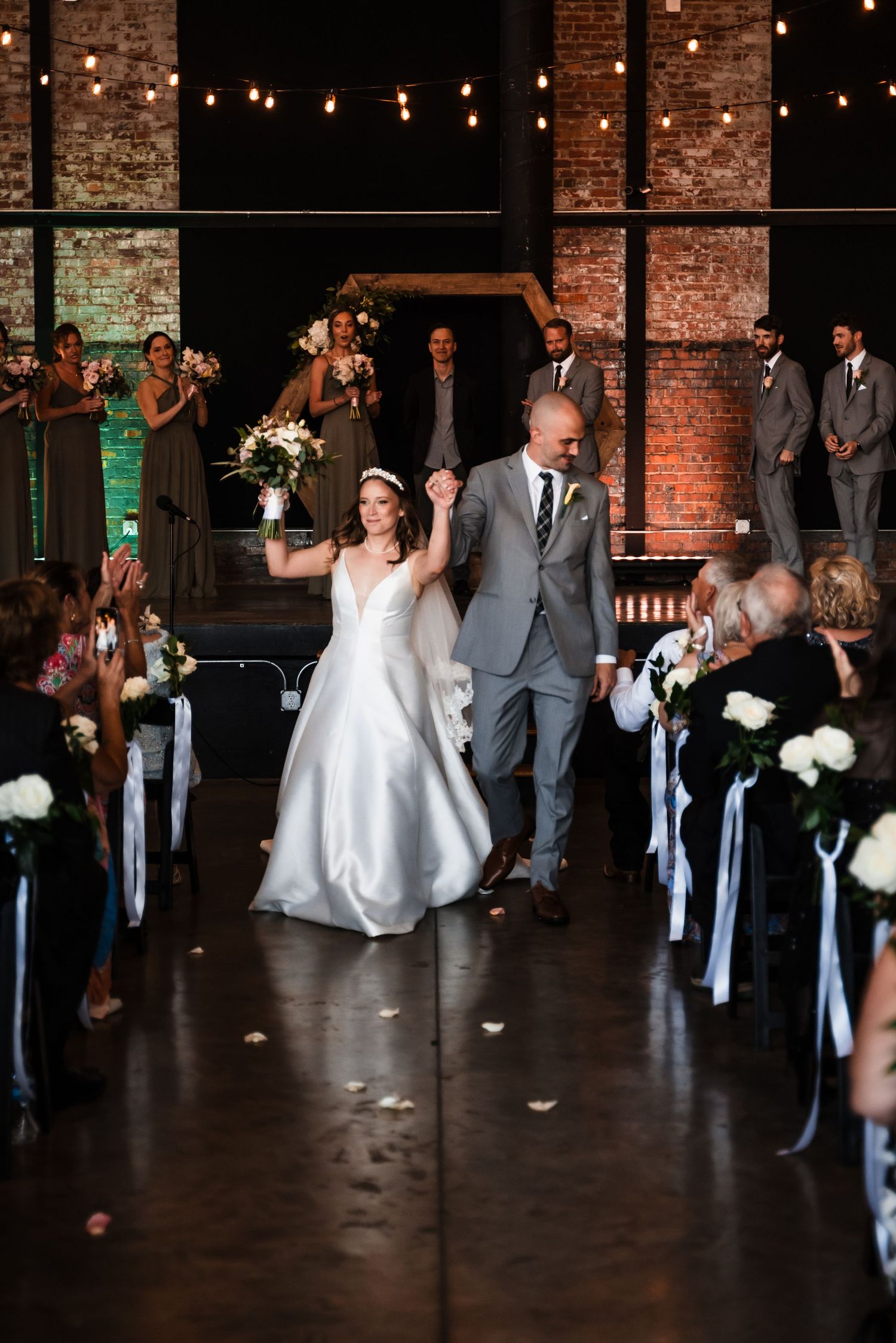 A happy couple celebrates while walking down the aisle at a wedding ceremony in a rustic brick venue with string lights.