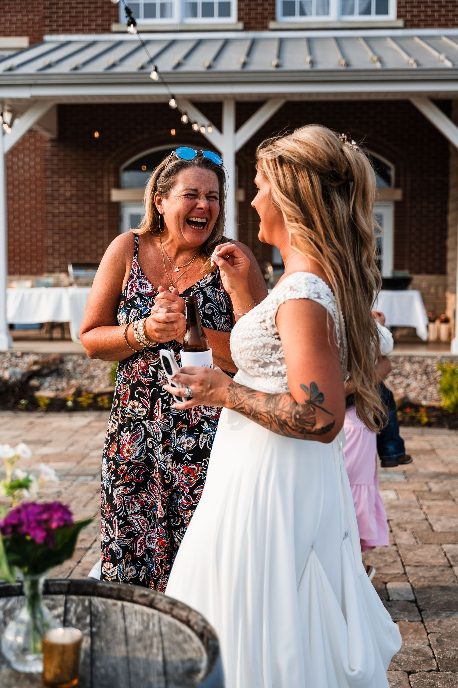 Two women sharing a joyful laugh during outdoor wedding reception.