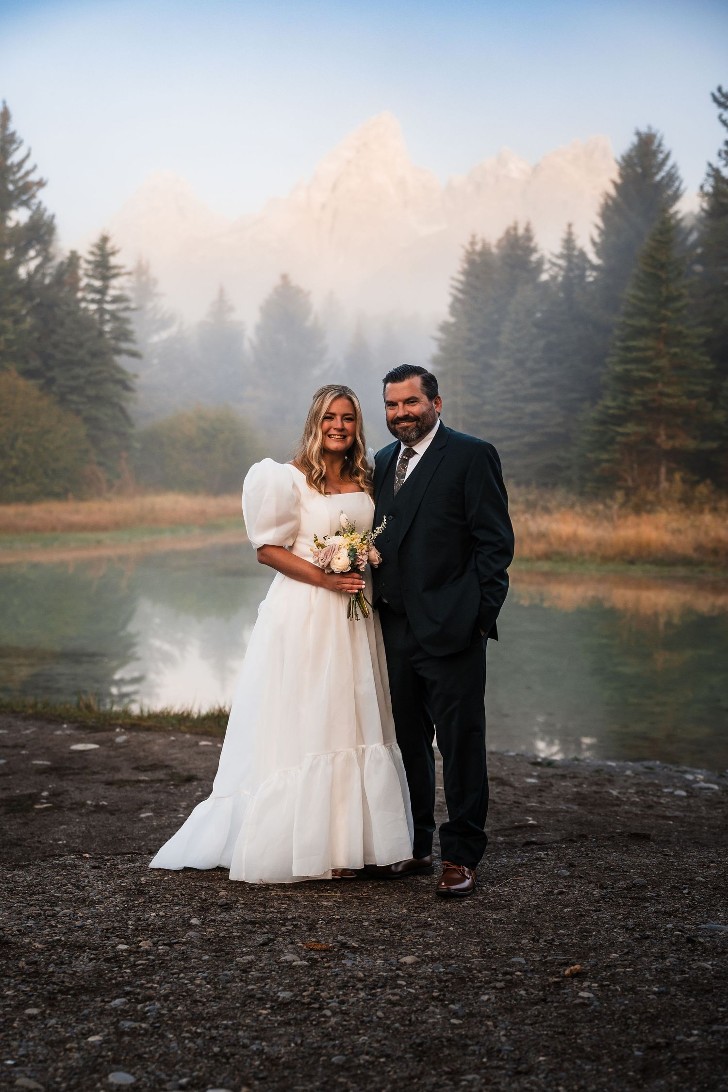 A couple poses for wedding photos by a tranquil lake during sunset.