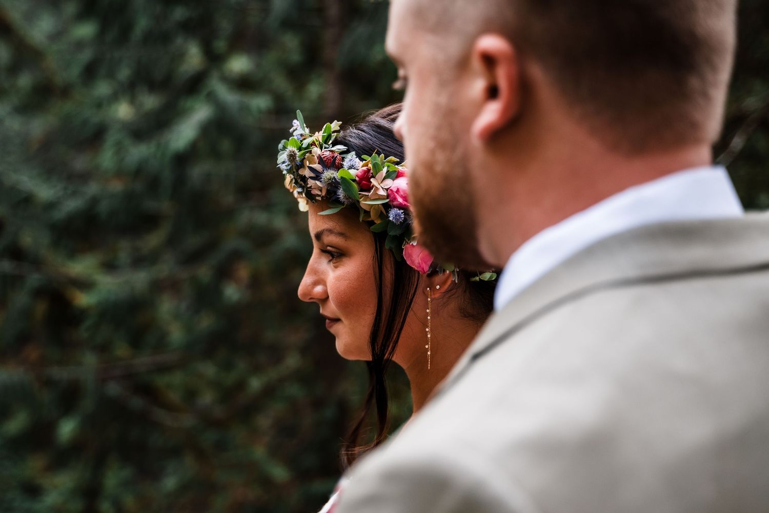 A bride wearing a delicate floral crown stands in profile next to a groom in a light gray suit during an outdoor ceremony.